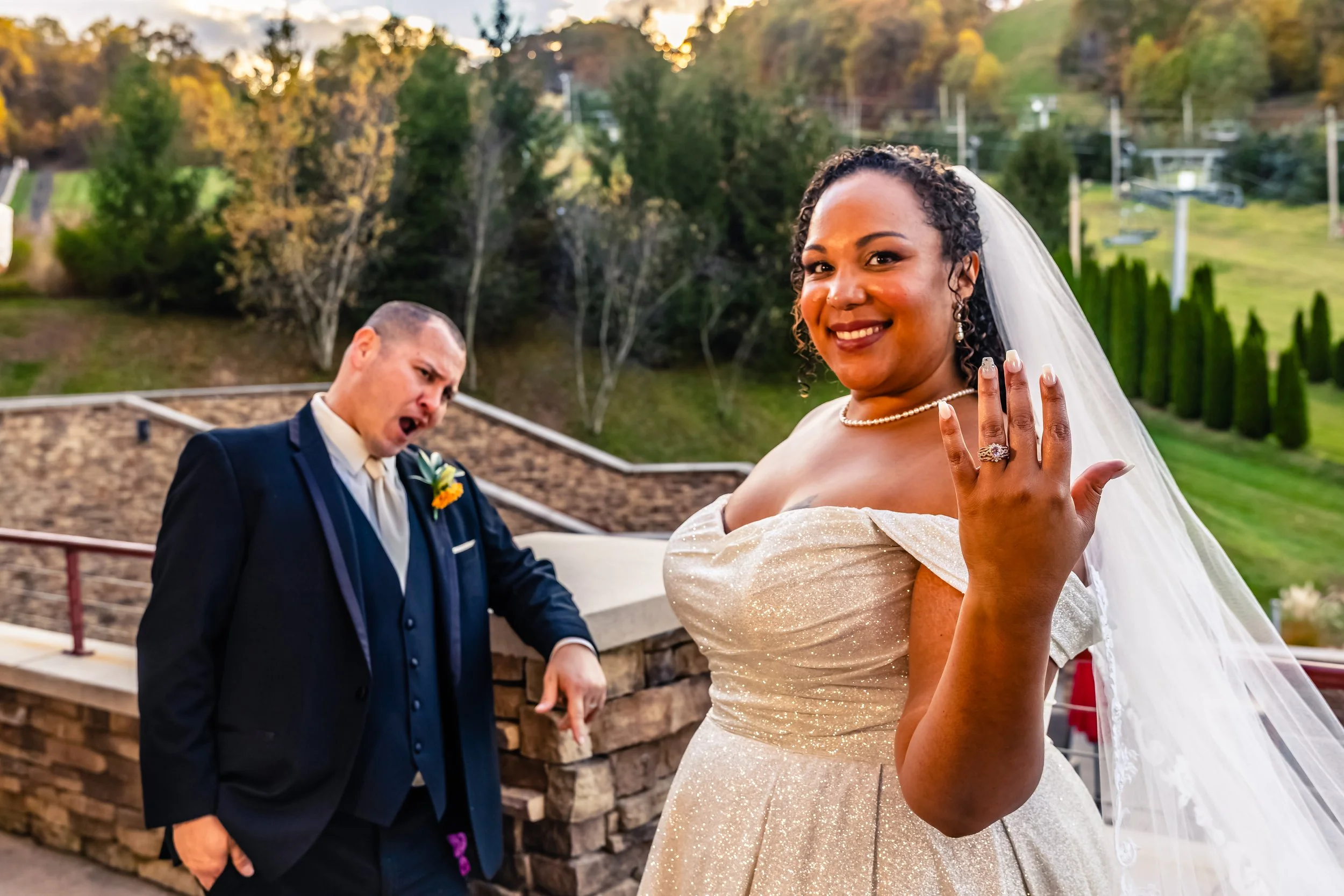 A bride showing her wedding ring, smiling at camera, with a groom in the background who appears surprised or excited. The scene is outdoors during sunset, with green trees and hills in the background.
