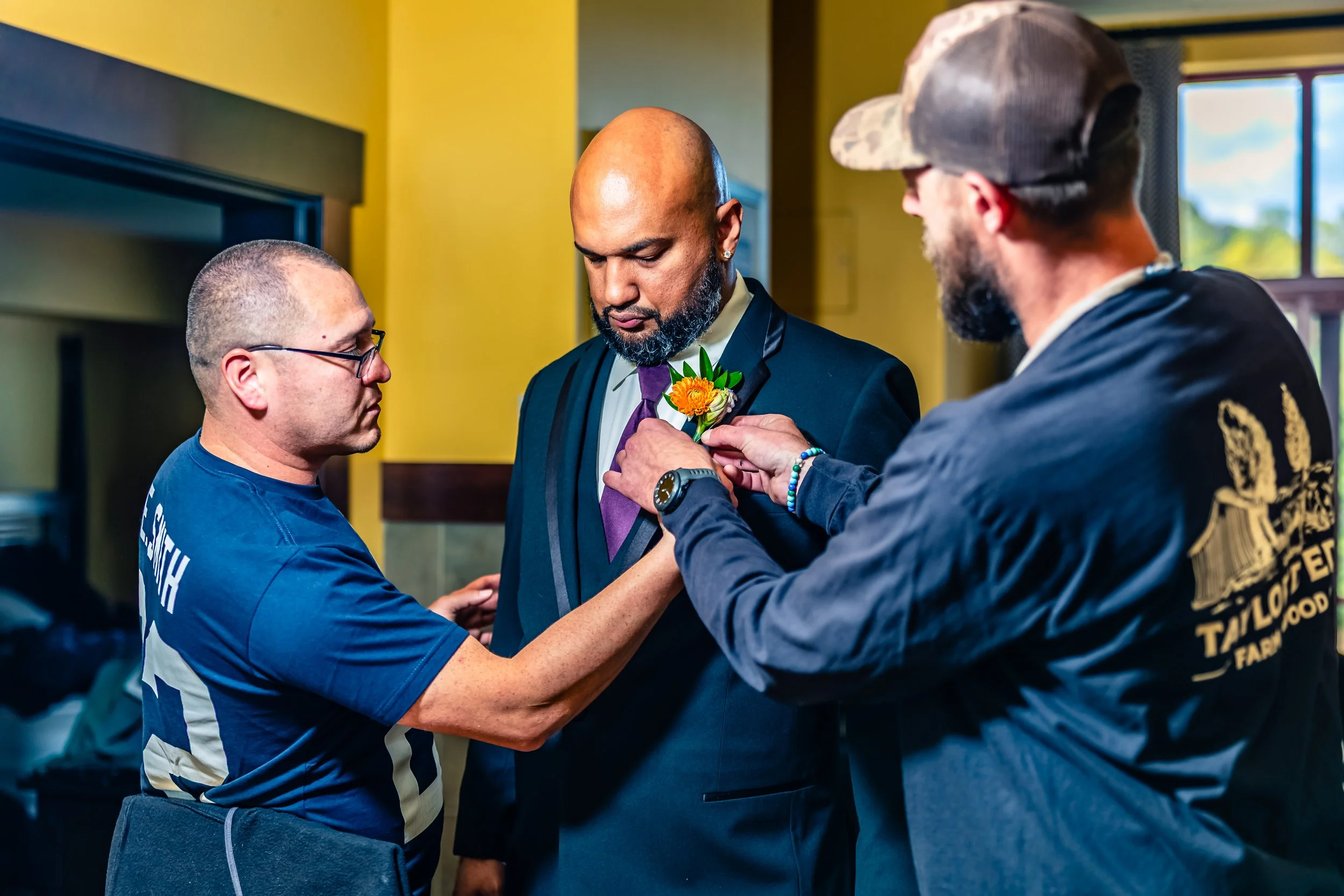 Two men pin a yellow flower boutonniere on a man in a tuxedo while another man assists. The man in the middle has a beard and is bald, wearing a black tuxedo with a purple tie, and the other two men are wearing shirts with logos.