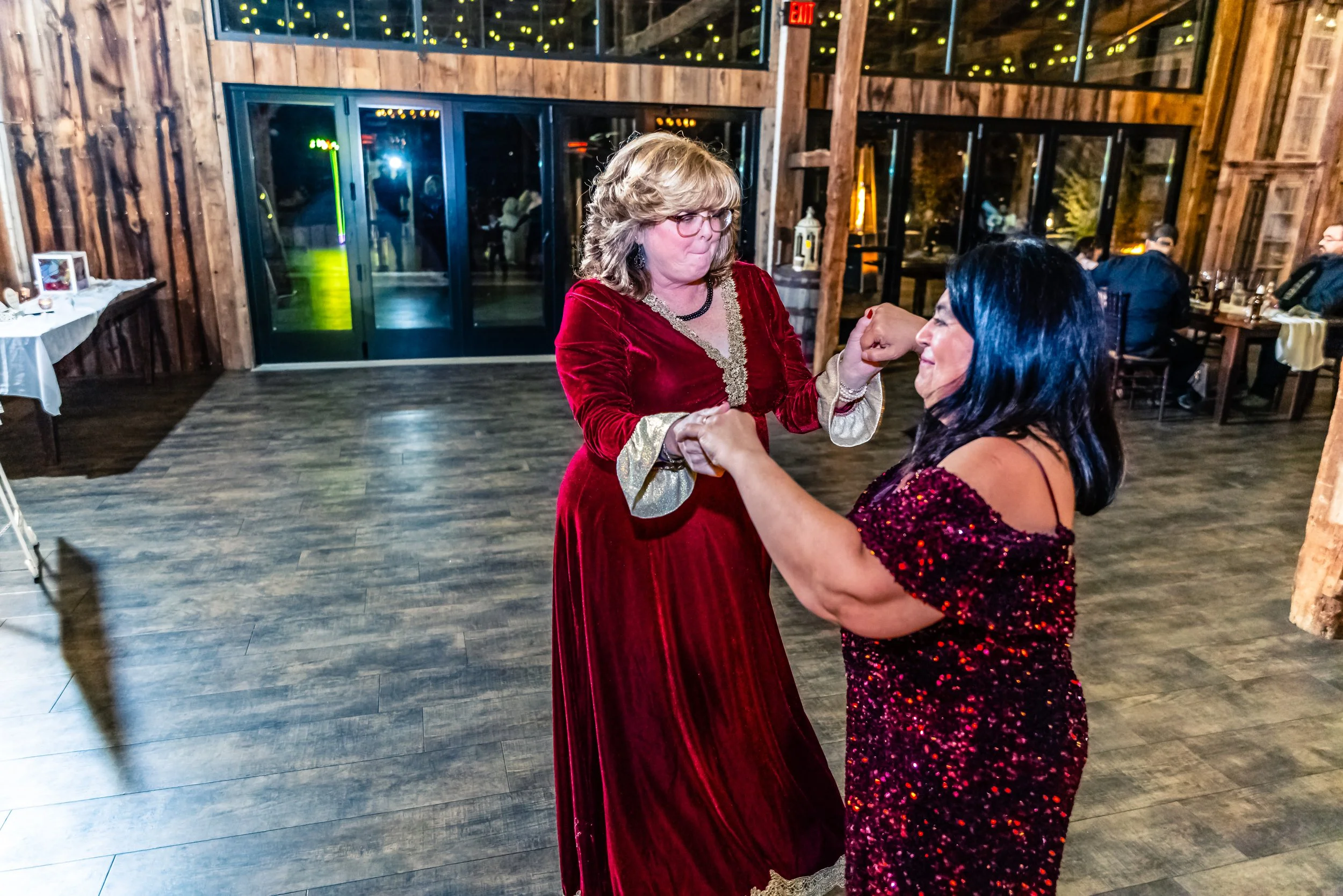 Two women dancing together at a celebration; one in a red velvet gown with gold accents, and the other in a sparkly red dress, inside a rustic wooden venue with tables and other people in the background.