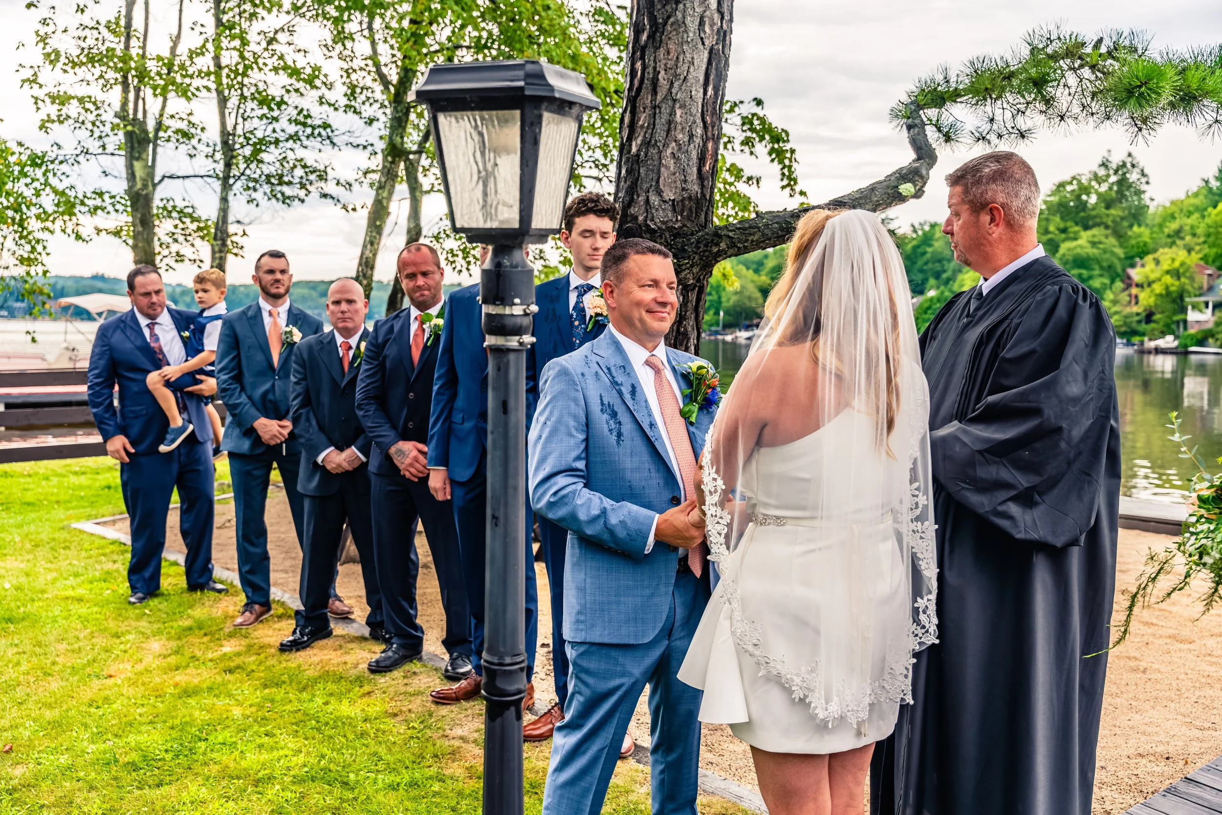 A wedding ceremony outdoors by a lake with a bride, groom, and officiant in focus, surrounded by groomsmen and a young boy, under a large tree with green foliage.