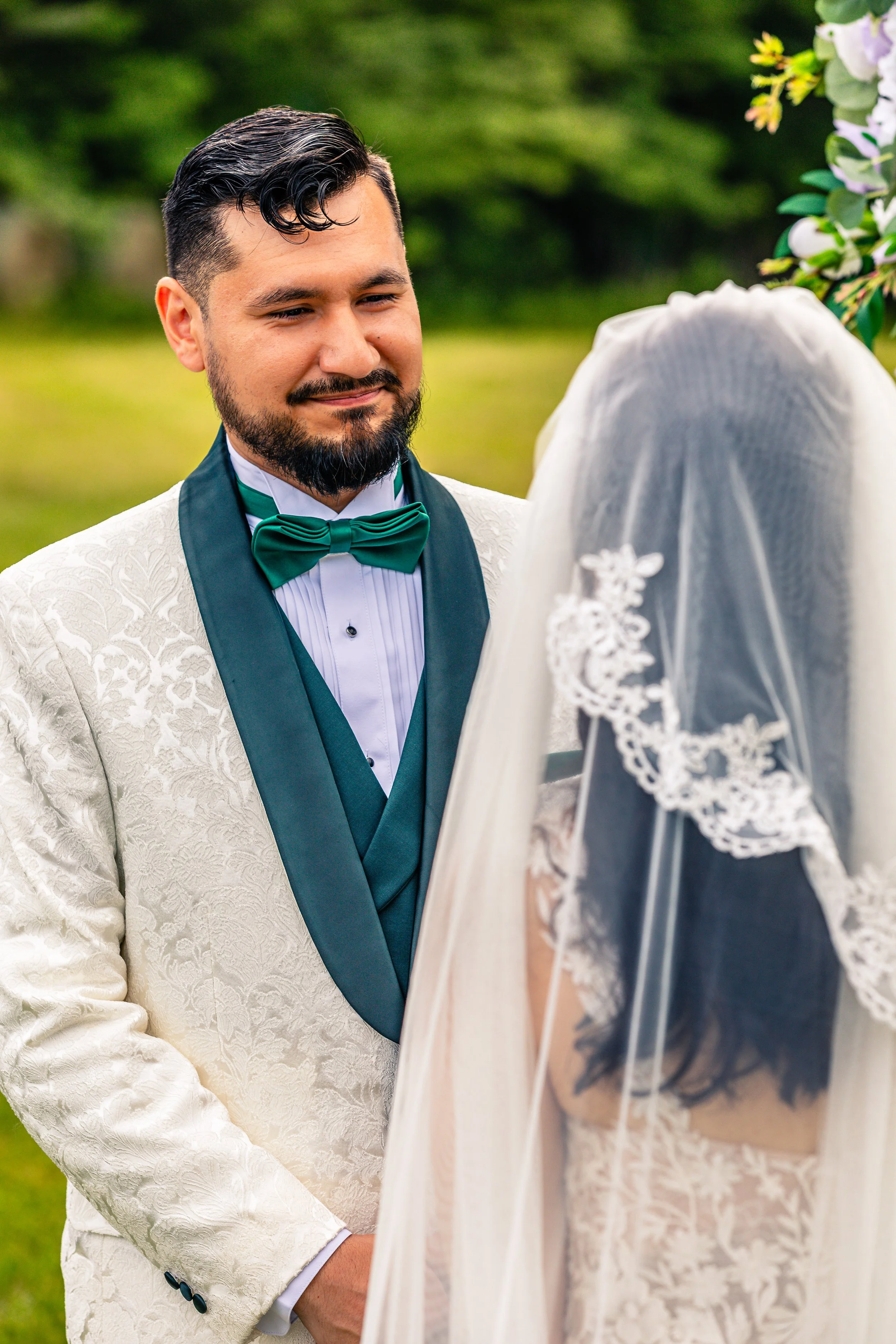A groom in a cream-colored, patterned tuxedo with a green bow tie and dark green sash, smiling and looking at a bride with a lace veil and wedding dress during an outdoor wedding ceremony.
