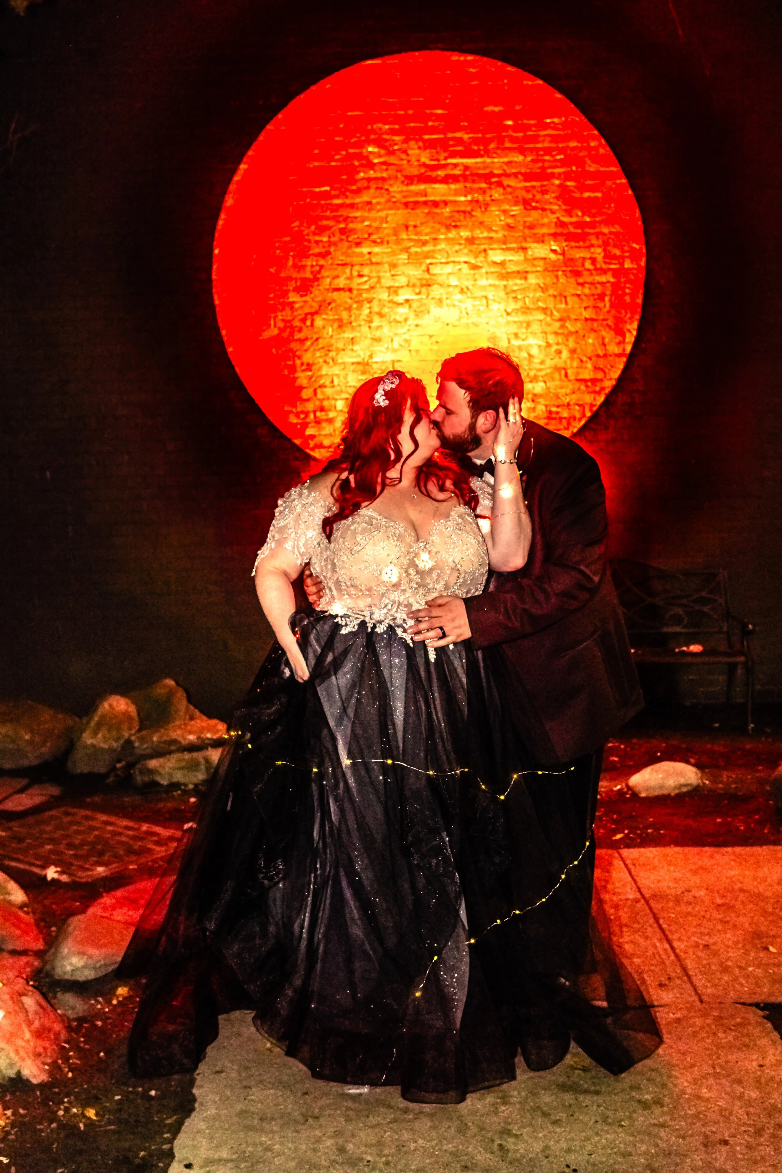 A couple sharing a kiss during their wedding, with a red-orange luminary on a dark wall behind them. The bride has red hair and is wearing a lace wedding dress, and the groom has dark hair and facial hair, dressed in a black suit.