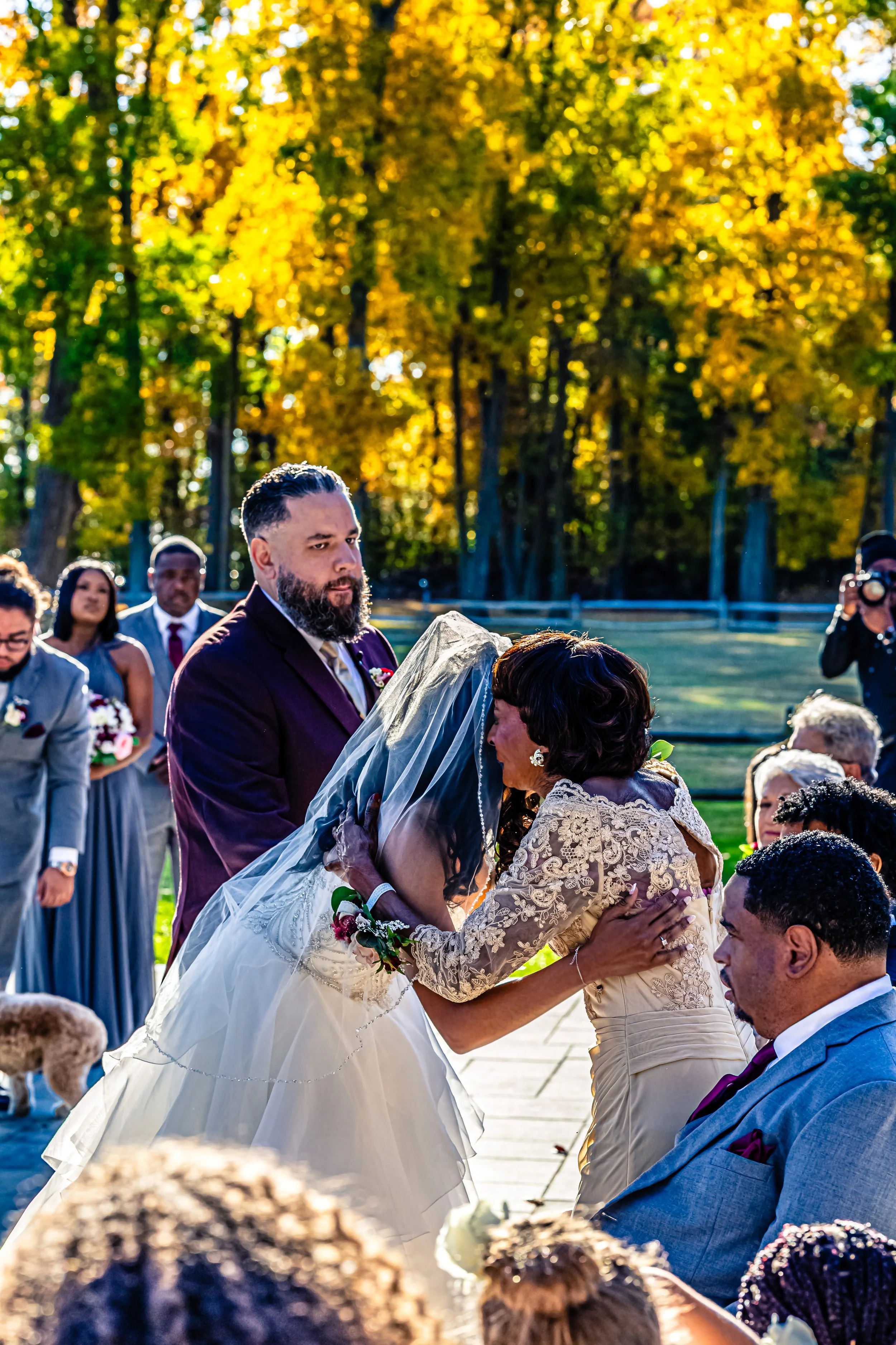 A wedding ceremony outdoors with a bride and groom exchanging vows, surrounded by family and friends, during fall with yellow and orange leaves in the background.