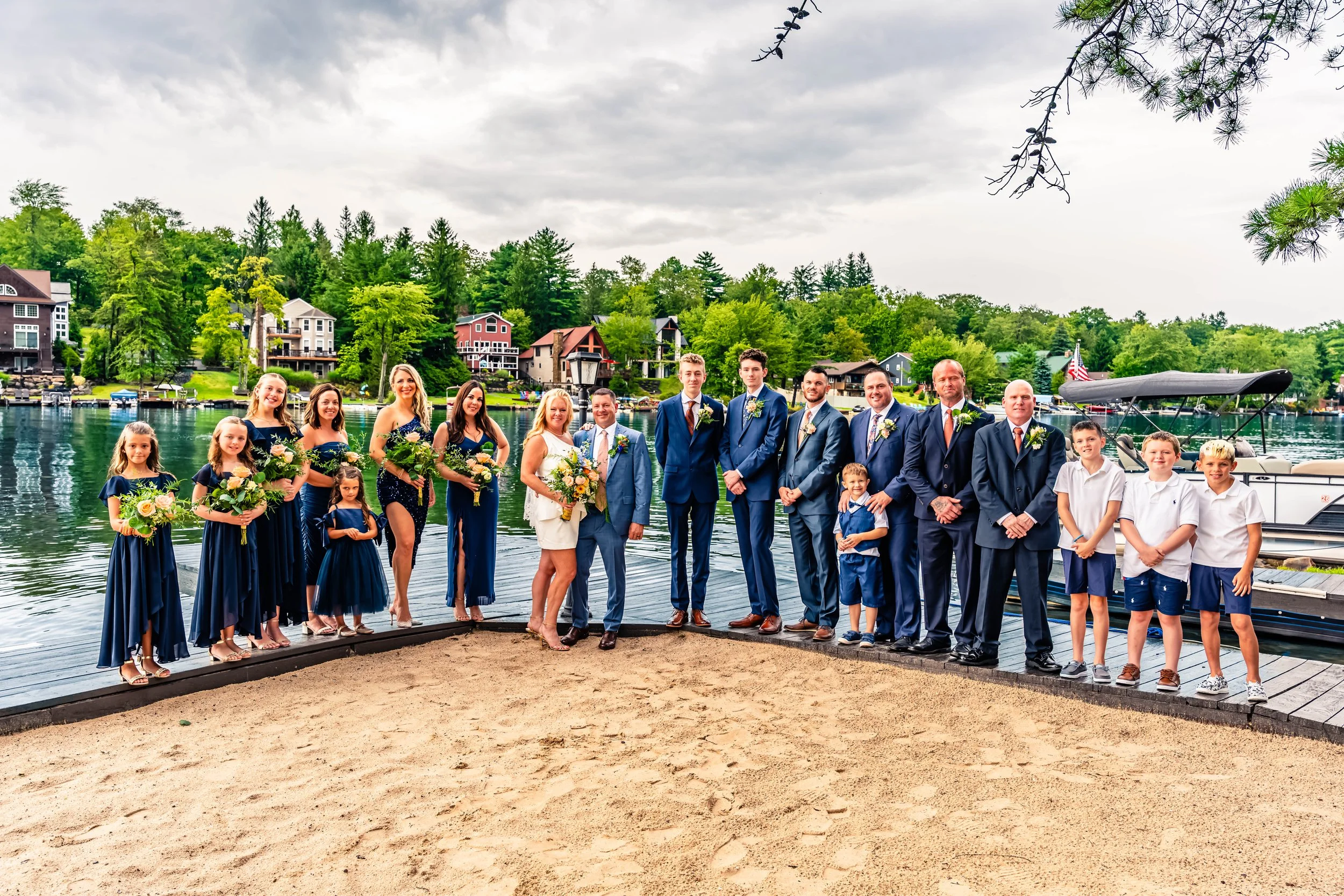 Group of people dressed in formal attire standing on a dock near a lake with houses and trees in the background during daytime.