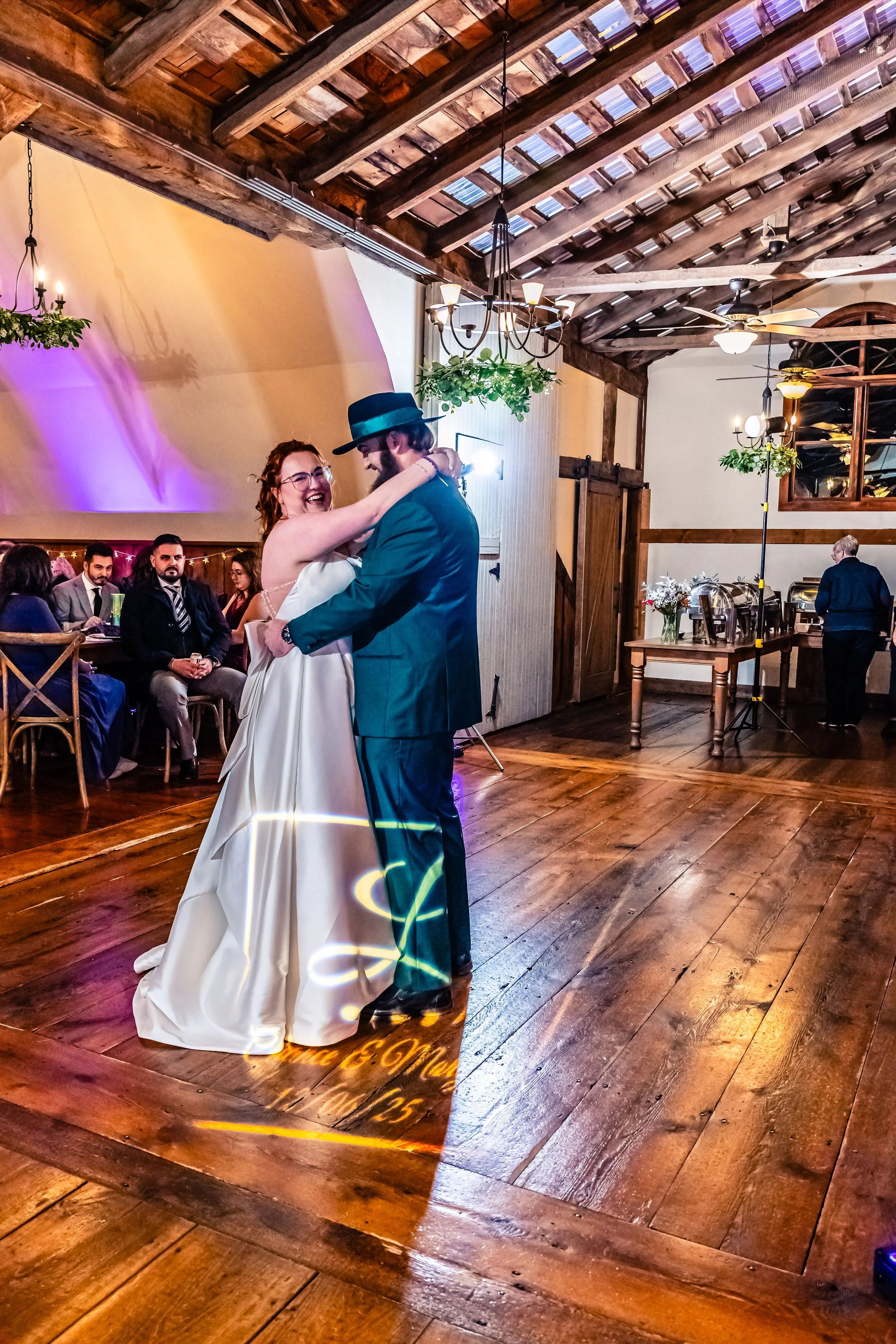 A bride and groom are dancing together at a wedding reception in a rustic barn with wooden floors, beams, and warm lighting.