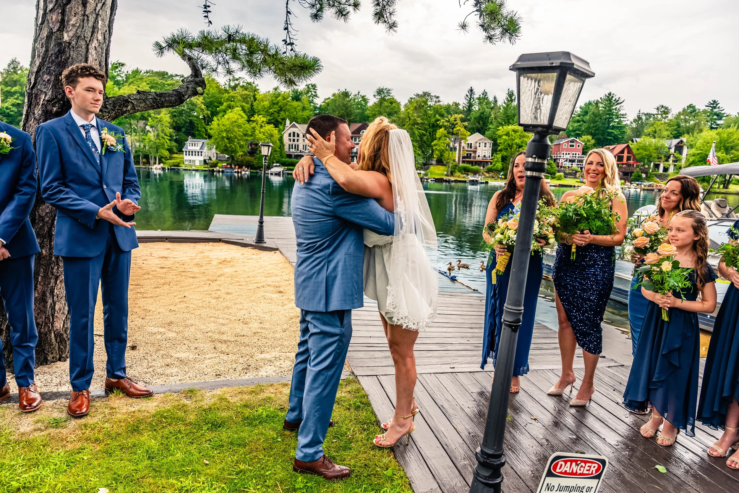 A wedding ceremony near a lake with trees and houses in the background, featuring a bride and groom embracing, surrounded by bridesmaids and groomsmen on a wooden dock and grassy area.