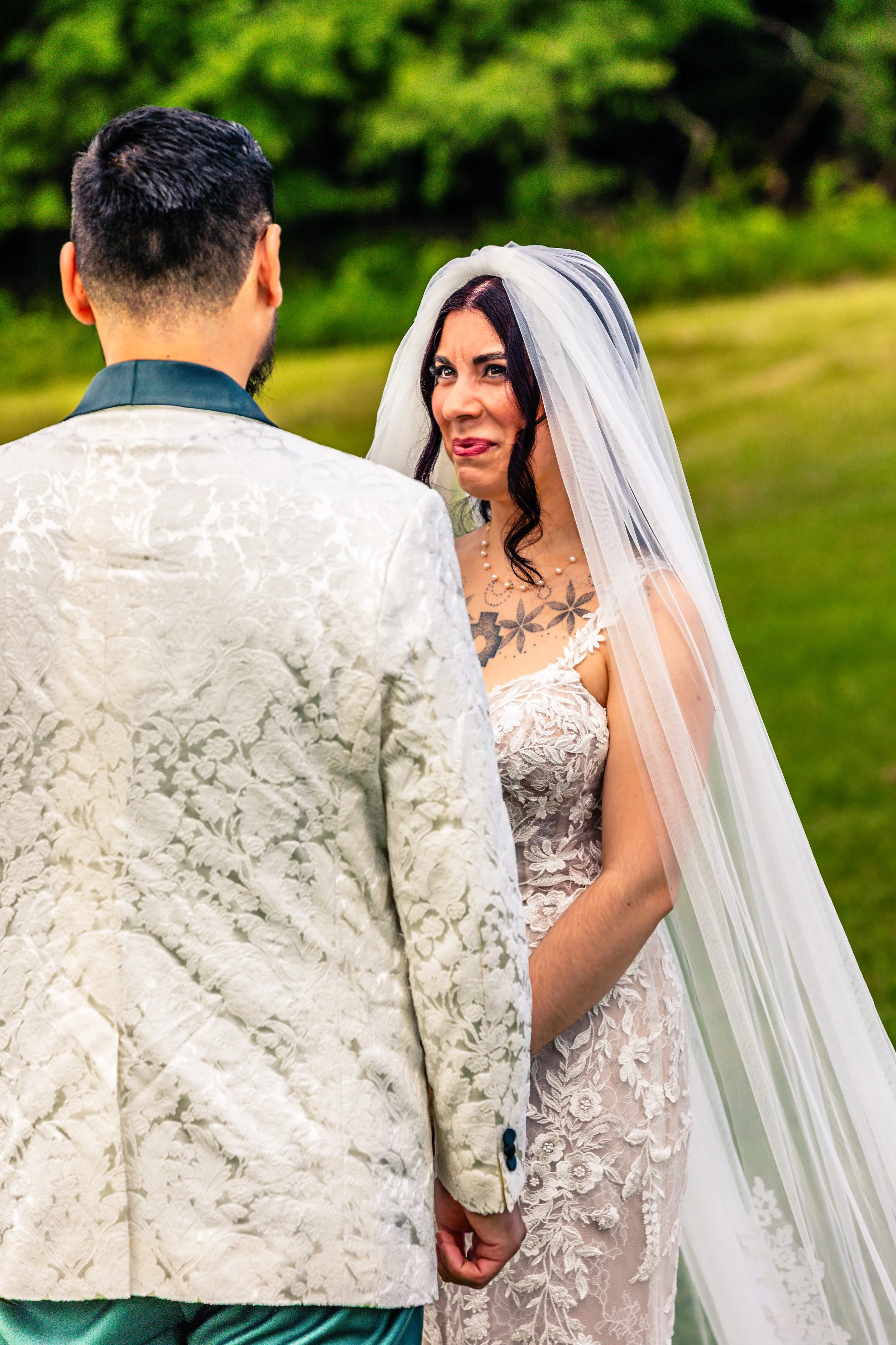 Bride and groom holding hands during their wedding ceremony outdoors, bride wearing a lace wedding dress with a long veil and the groom wearing a white floral patterned jacket.