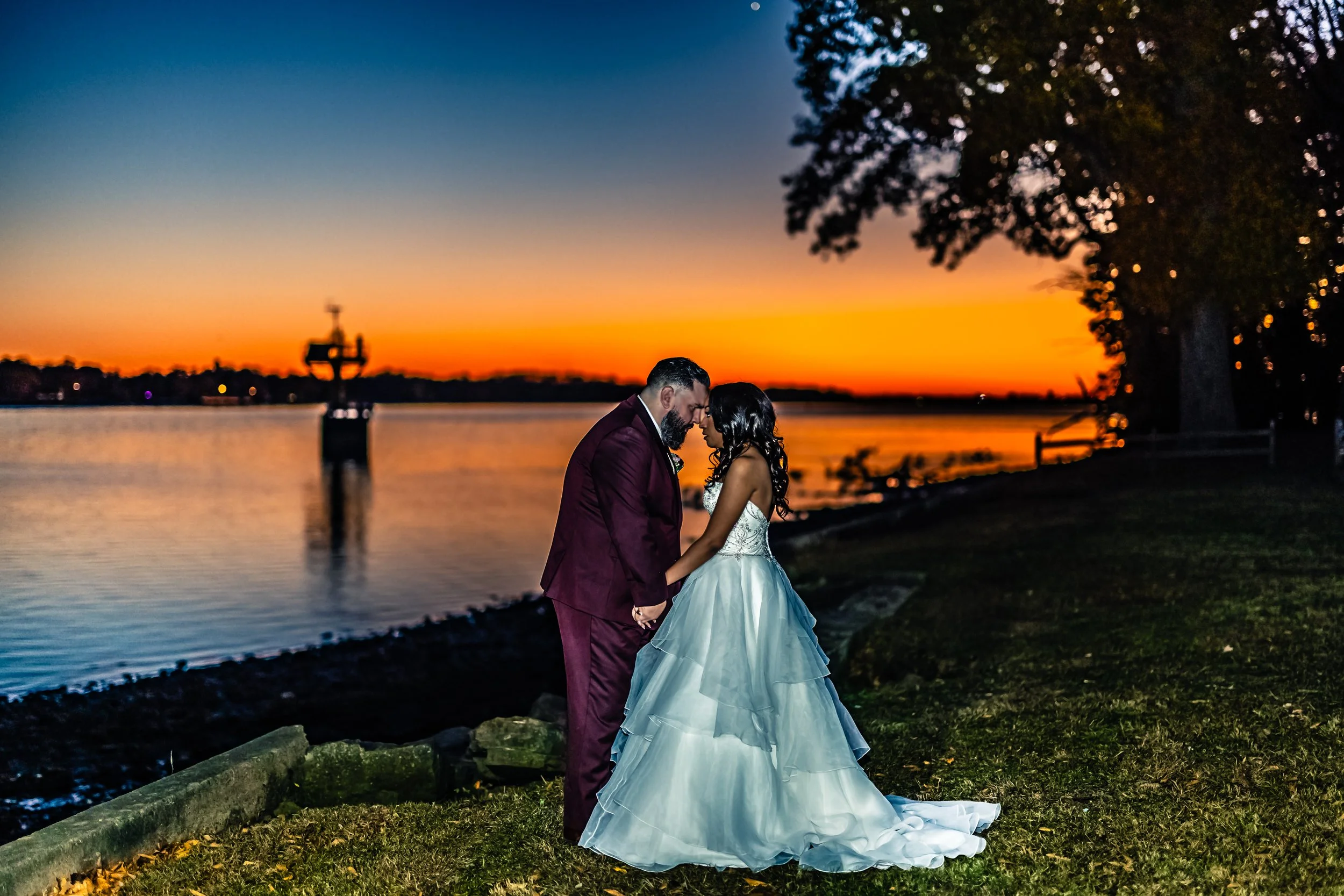 A bride and groom stand close together, holding hands with foreheads touching, outdoors near a body of water during sunset.