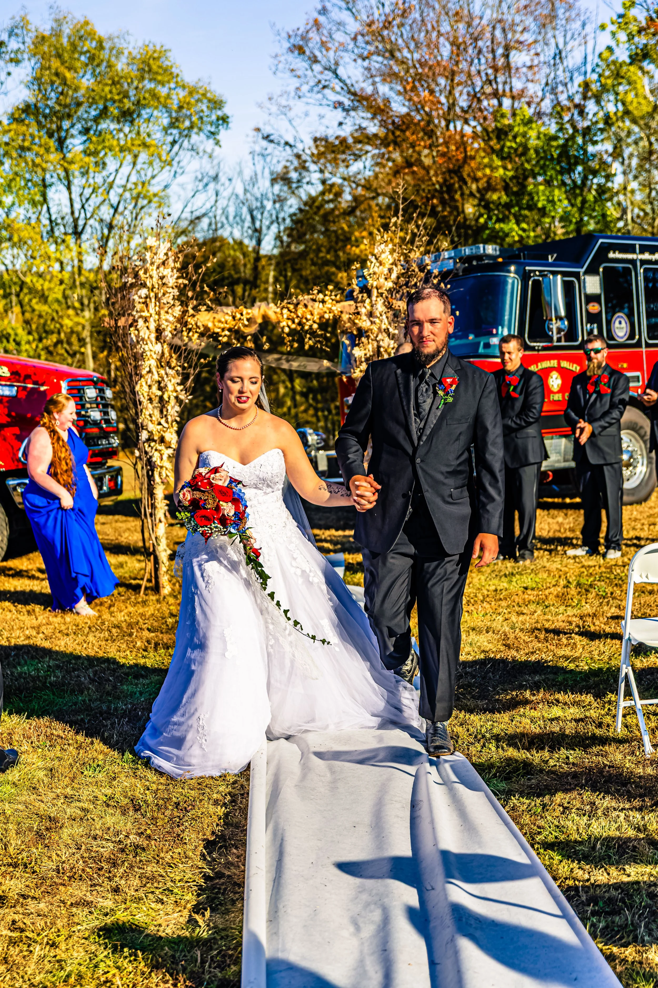 A bride and groom walk hand in hand down an outdoor aisle at a wedding ceremony, surrounded by fire trucks and trees with fall foliage in the background, on a sunny day.