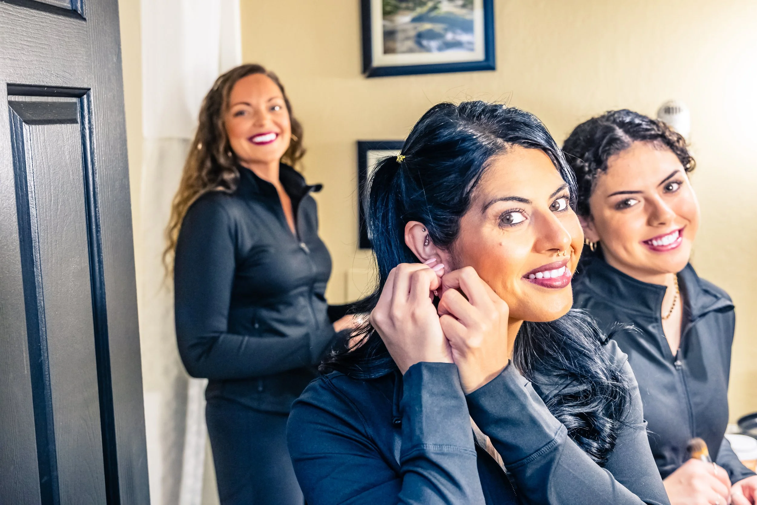 Three women wearing black jackets smiling indoors, one is adjusting an earring, another is holding a makeup brush, and one is smiling in the background.