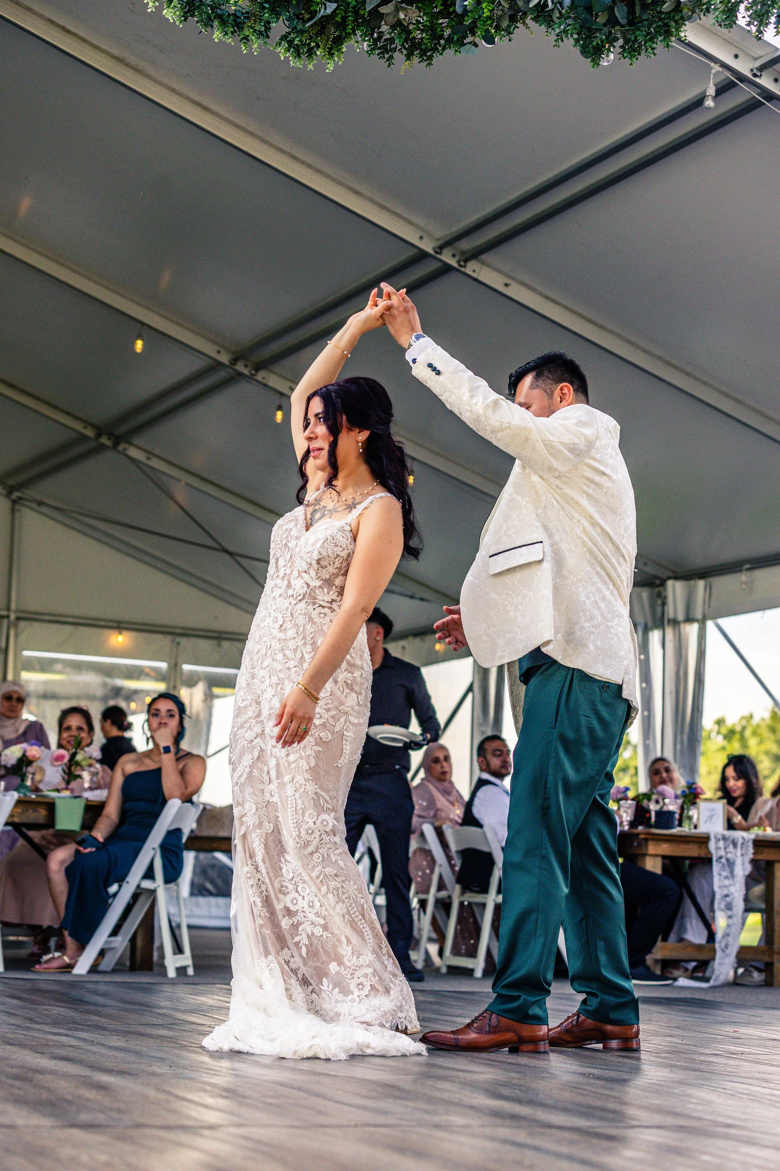 A bride and groom dancing at their wedding reception under a large tent, with guests sitting at decorated tables watching.