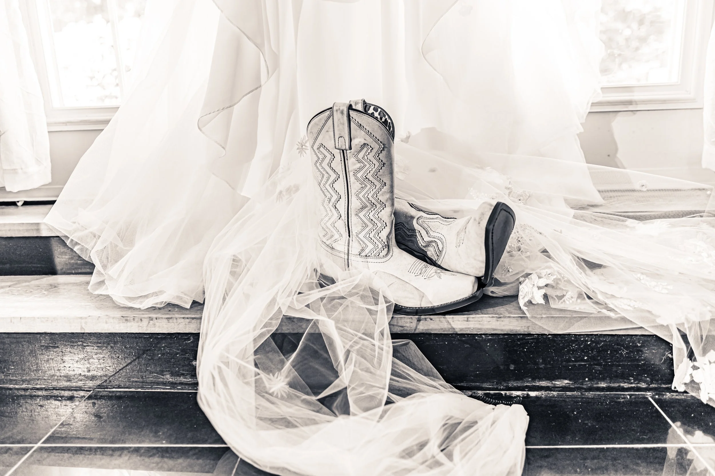 A pair of cowboy boots on a wooden floor, surrounded by sheer tulle fabric and a white bridal veil, with curtains and a window in the background.