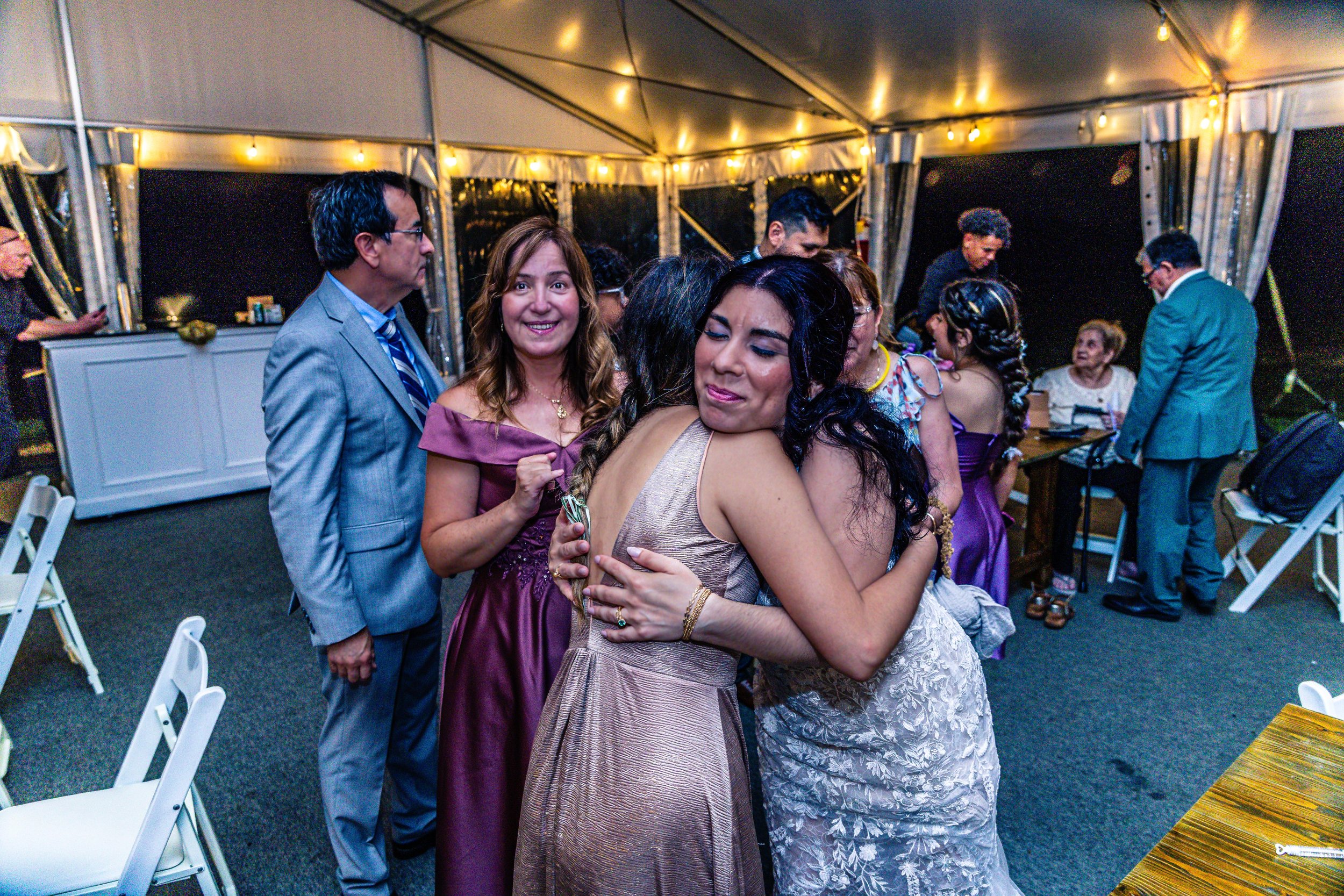Two women hugging at a celebration with people dancing and socializing in the background inside a decorated tent.