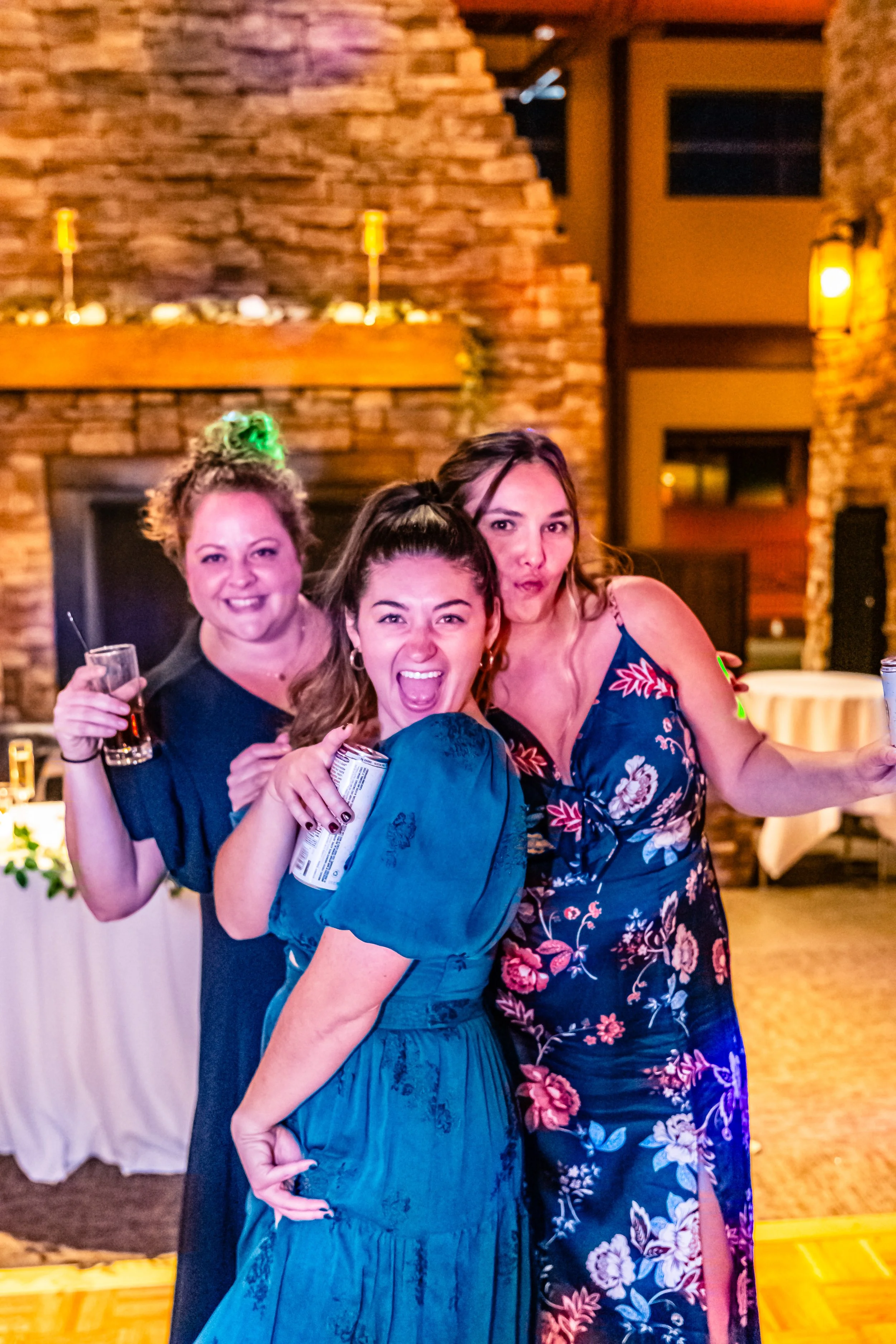 Three women at a party smiling and holding drinks, standing in a cozy room with a stone fireplace and warm lighting.