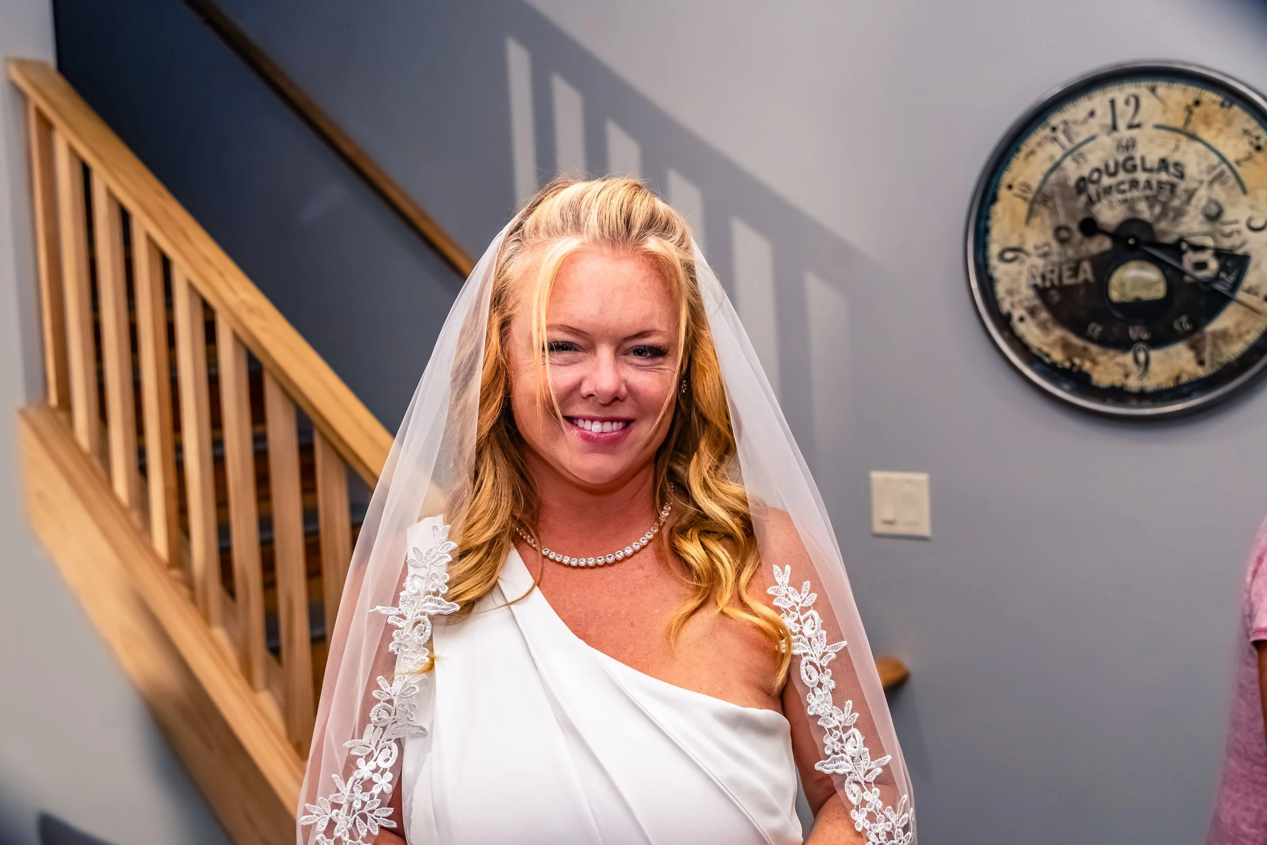 A smiling bride with blonde hair in a white dress, wearing a veil and pearl necklace, standing indoors near a staircase.