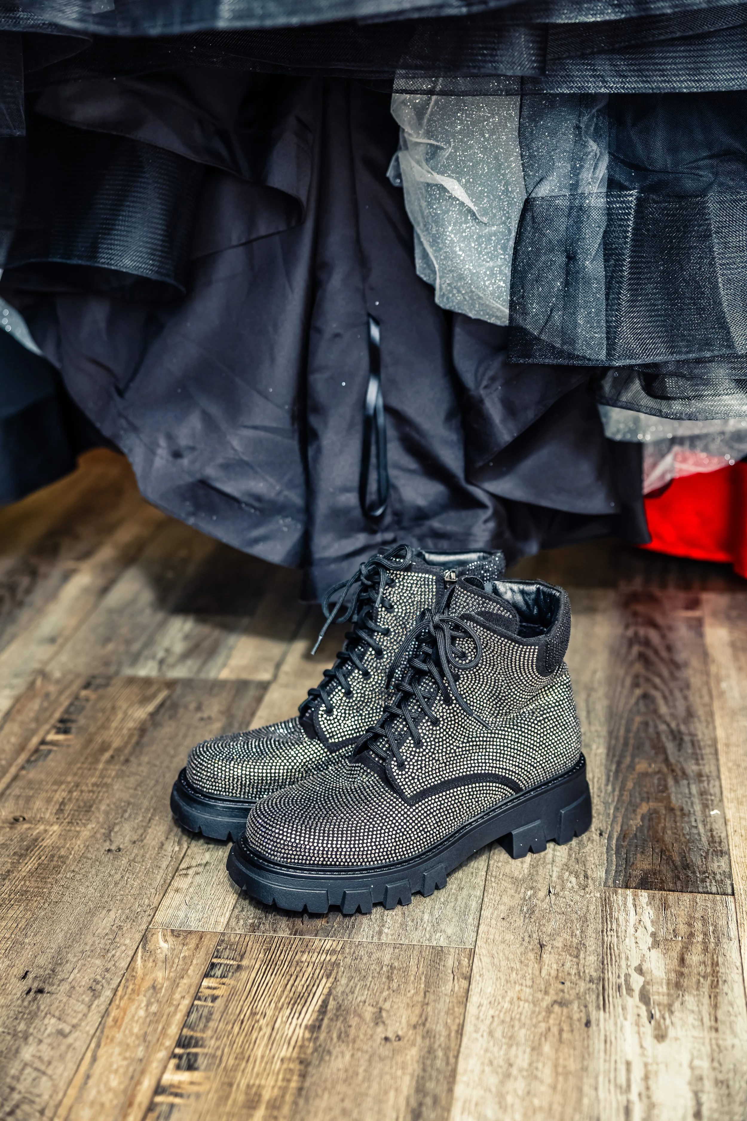 Pair of sparkly, black and silver combat boots on a wooden floor in front of a black and decorated dress or skirt.