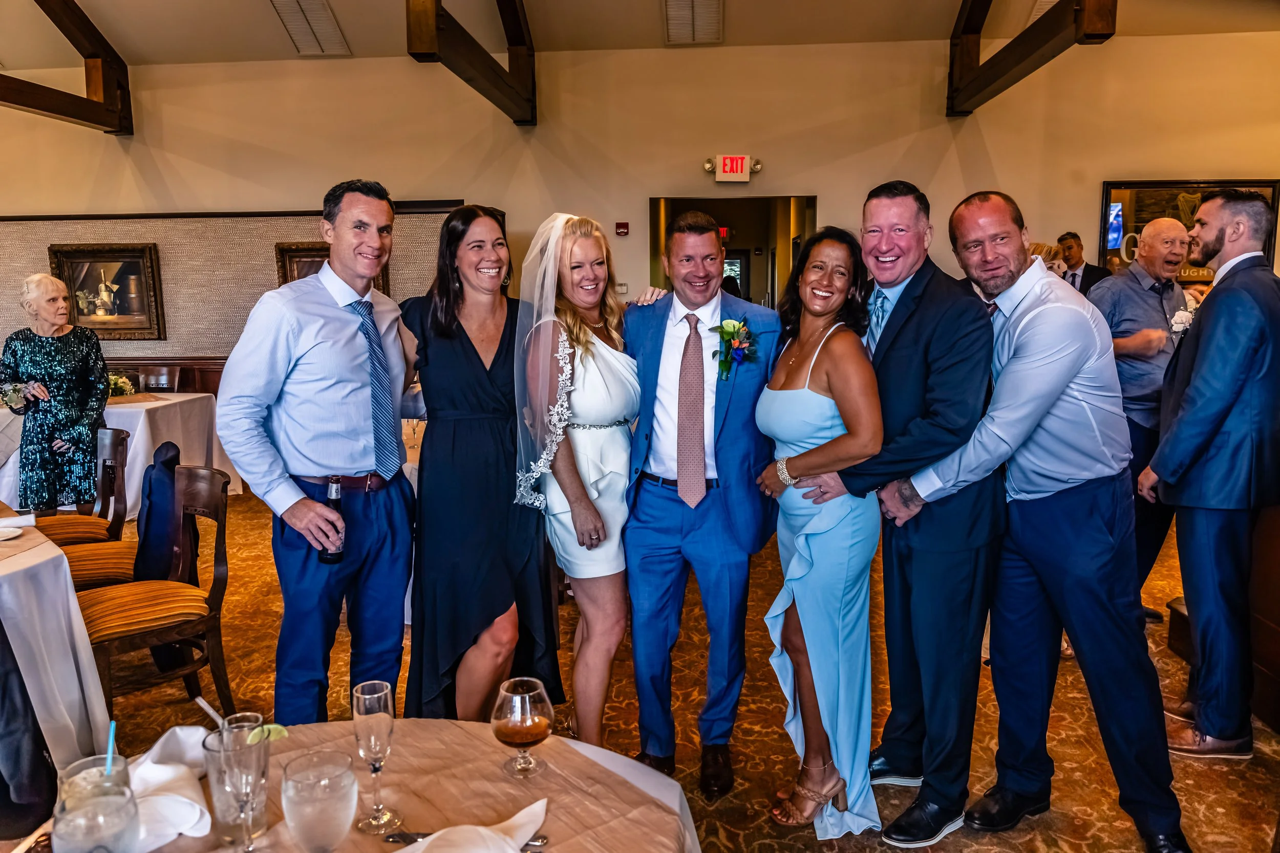 A group of nine people celebrating at a wedding reception, dressed in formal attire, smiling and posing together.