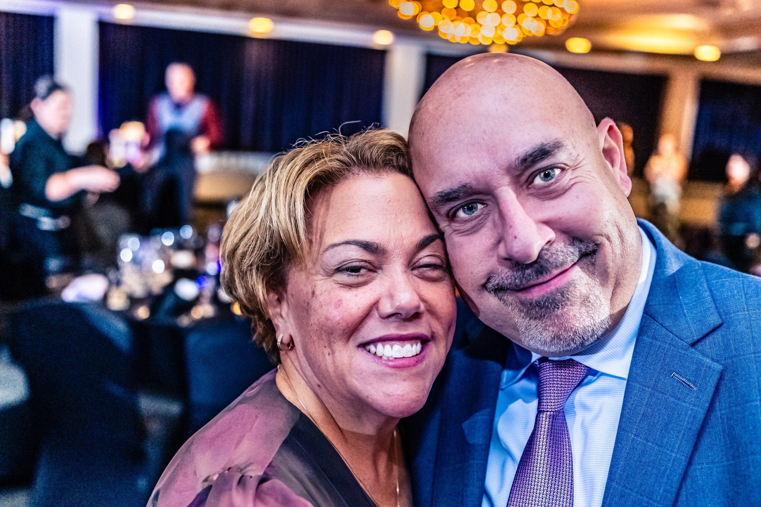 Close-up of a smiling woman and man in formal attire at a social event, with a blurred background of other people and decorations.