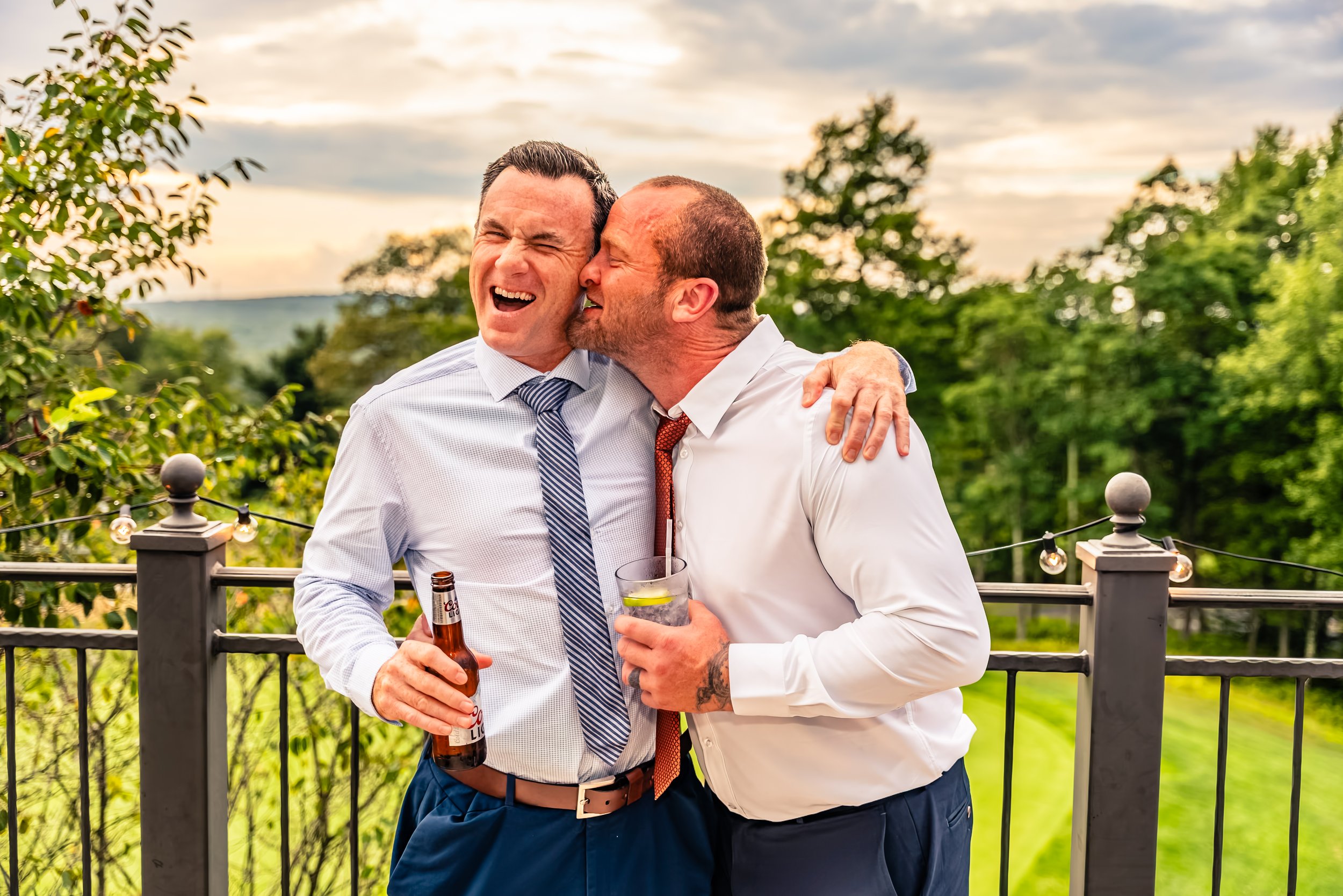 Two men in formal clothing sharing a moment on a balcony; one has his arm around the other, holding a beer, while the other is kissing him on the cheek, both smiling and appearing joyful, with a background of green trees and a cloudy sky.