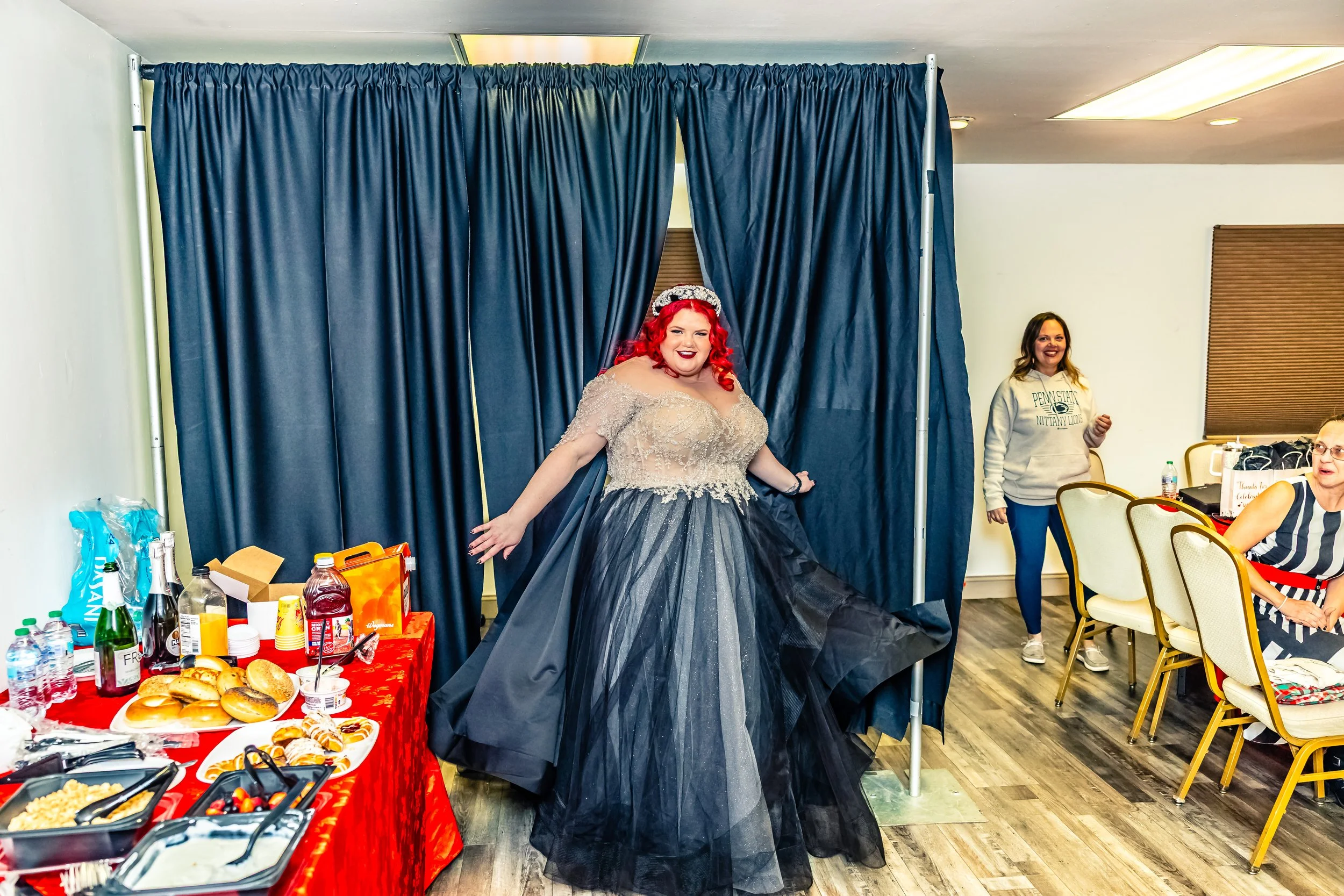 A woman with bright red hair in an elegant gown stands on a small stage with a dark blue curtain, smiling and posing for the camera. There is a table with food and drinks on the left, and two women sitting and standing nearby in a room with wooden fl
