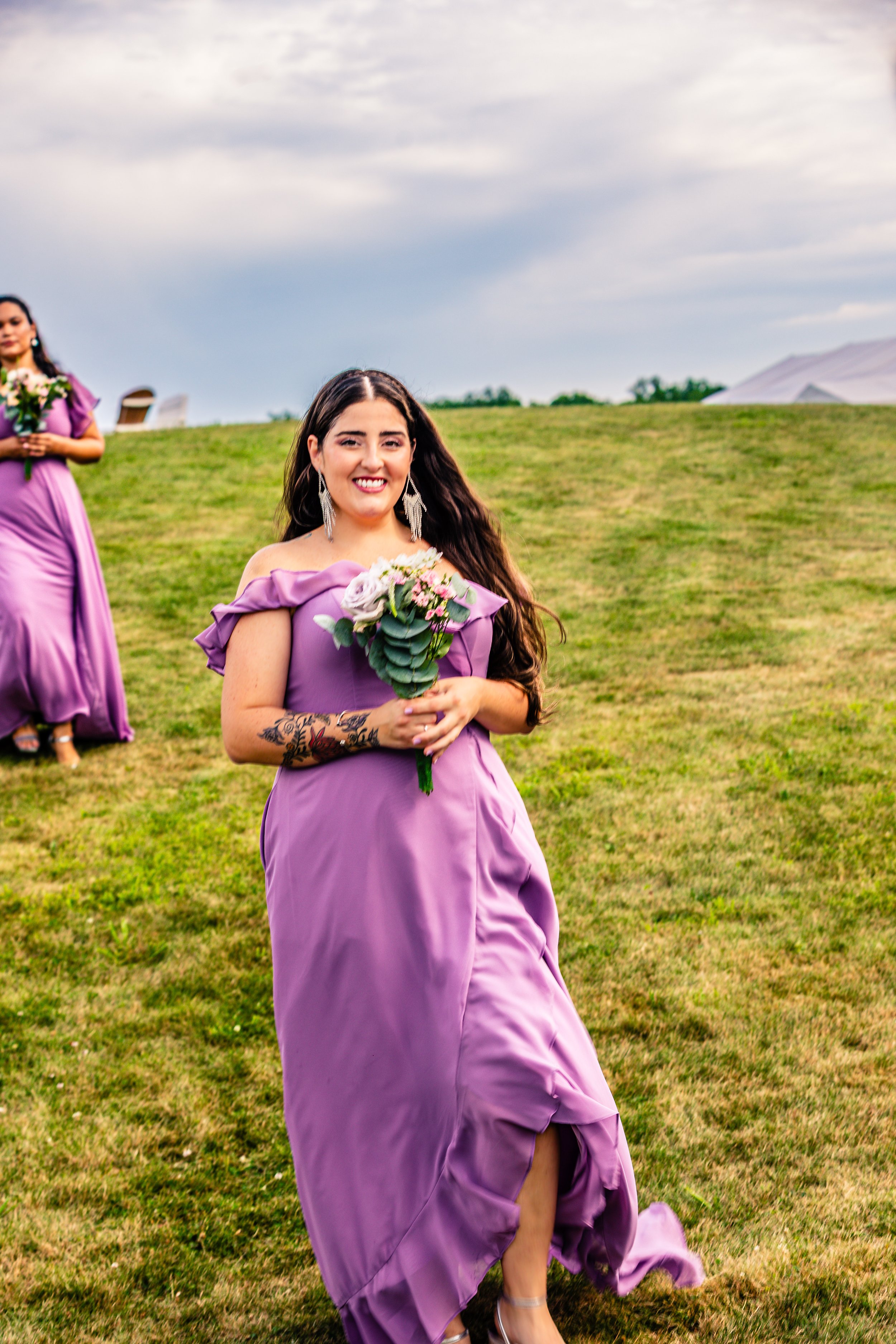 A woman in a purple dress holding a bouquet of flowers, smiling outdoors on a grassy field during the daytime. Another woman in a matching purple dress is visible in the background.