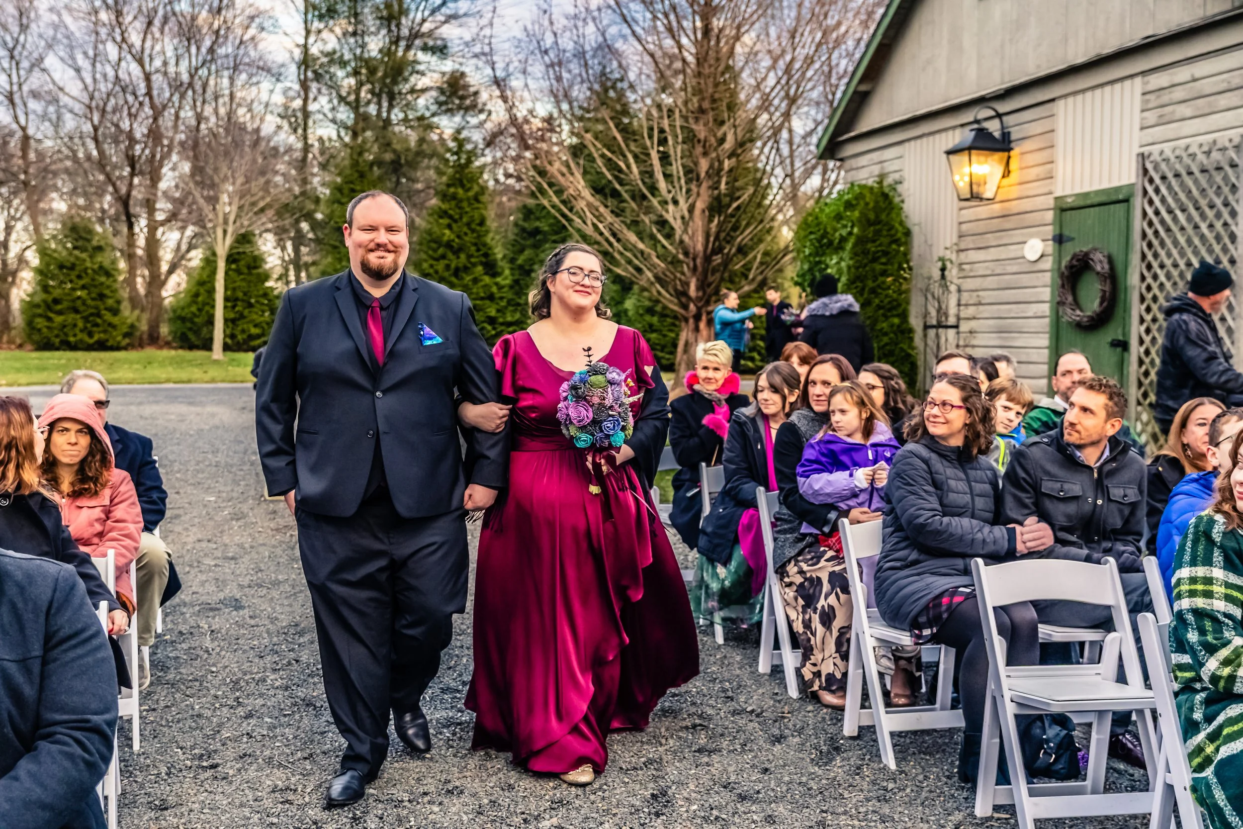 A wedding ceremony outdoors with a couple walking down the aisle, the bride holding a bouquet of purple and blue flowers, and guests seated on white folding chairs watching and smiling.
