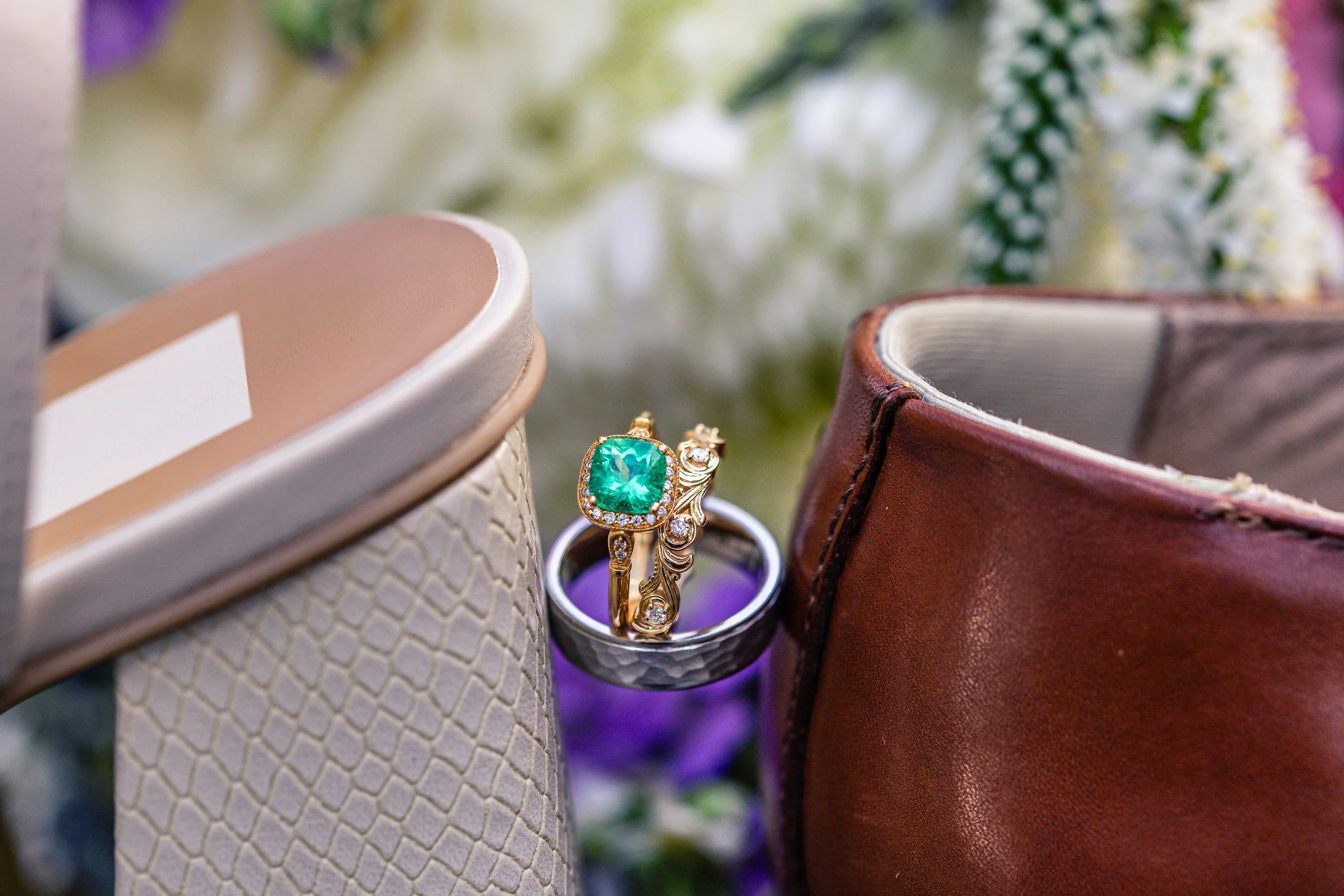 Close-up of a ring with an emerald center stone, surrounded by small diamonds, nestled between a pair of shoes and a brown leather bag, with white and purple flowers in the background.
