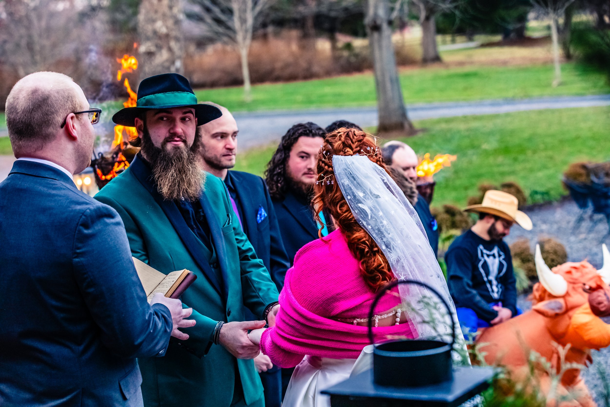 Couple getting married outdoors, with one person wearing a white veil and pink shawl, holding hands with officiant, surrounded by guests, and a costume buffalo in the background.
