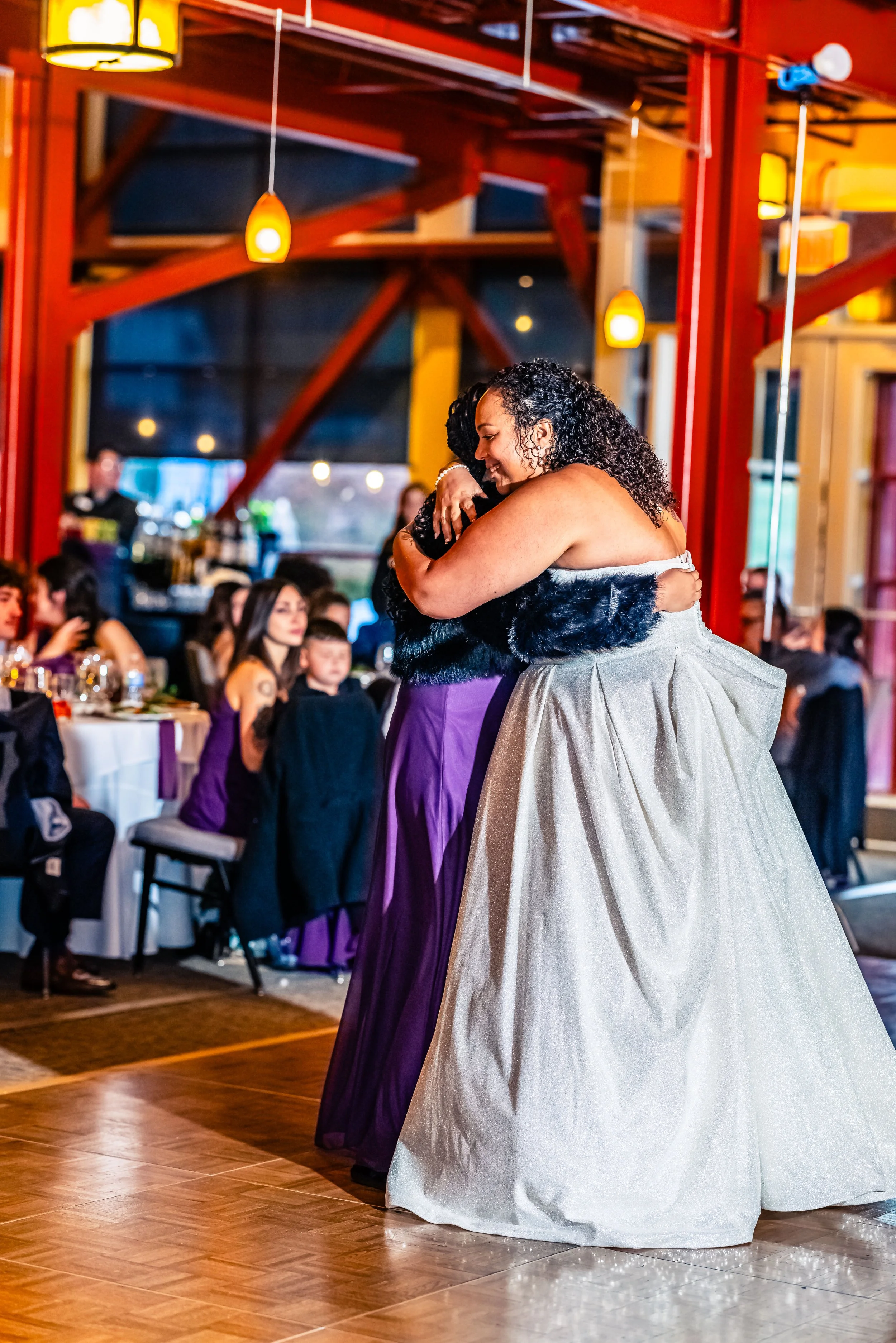 Two women hugging on a dance floor at a wedding reception, with guests seated at tables in the background.