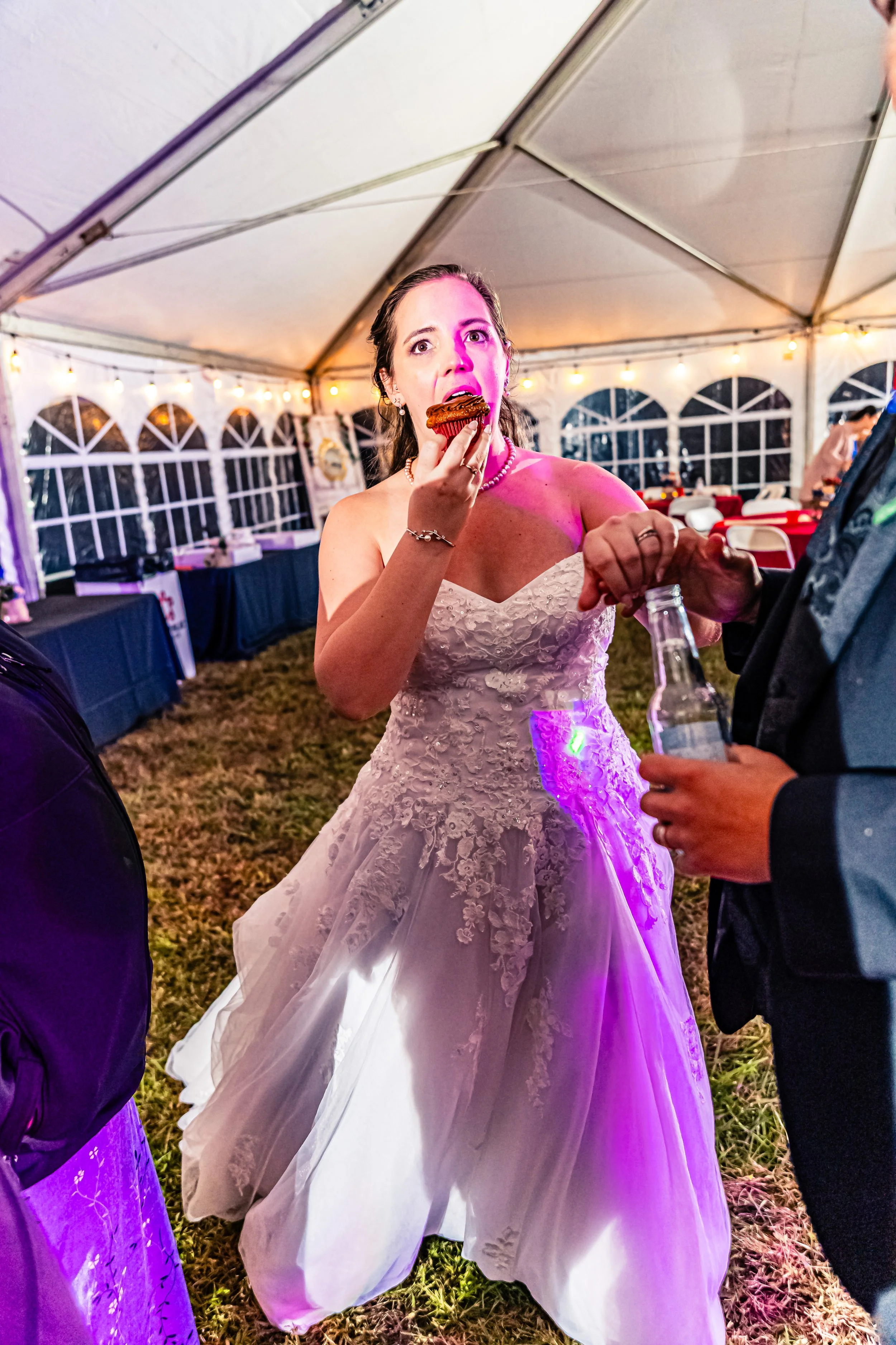 A bride in a white wedding gown and pearl necklace enjoys a cupcake at her wedding reception inside a large white tent decorated with string lights.
