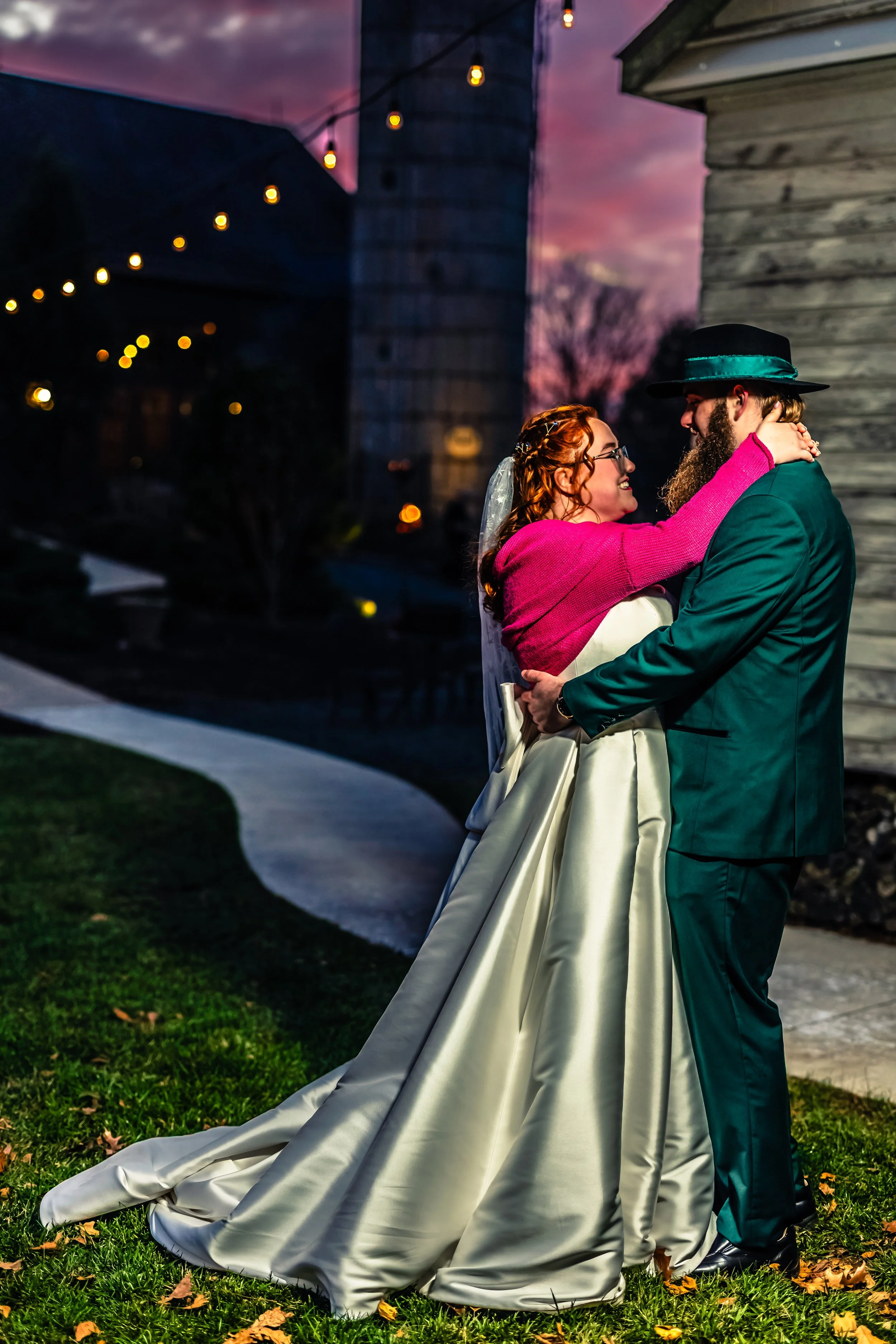 A bride and groom share a romantic dance outdoors at sunset, with string lights in the background. The bride is wearing a white wedding gown and a veil, and the groom is in a green suit with a black hat featuring a teal ribbon.