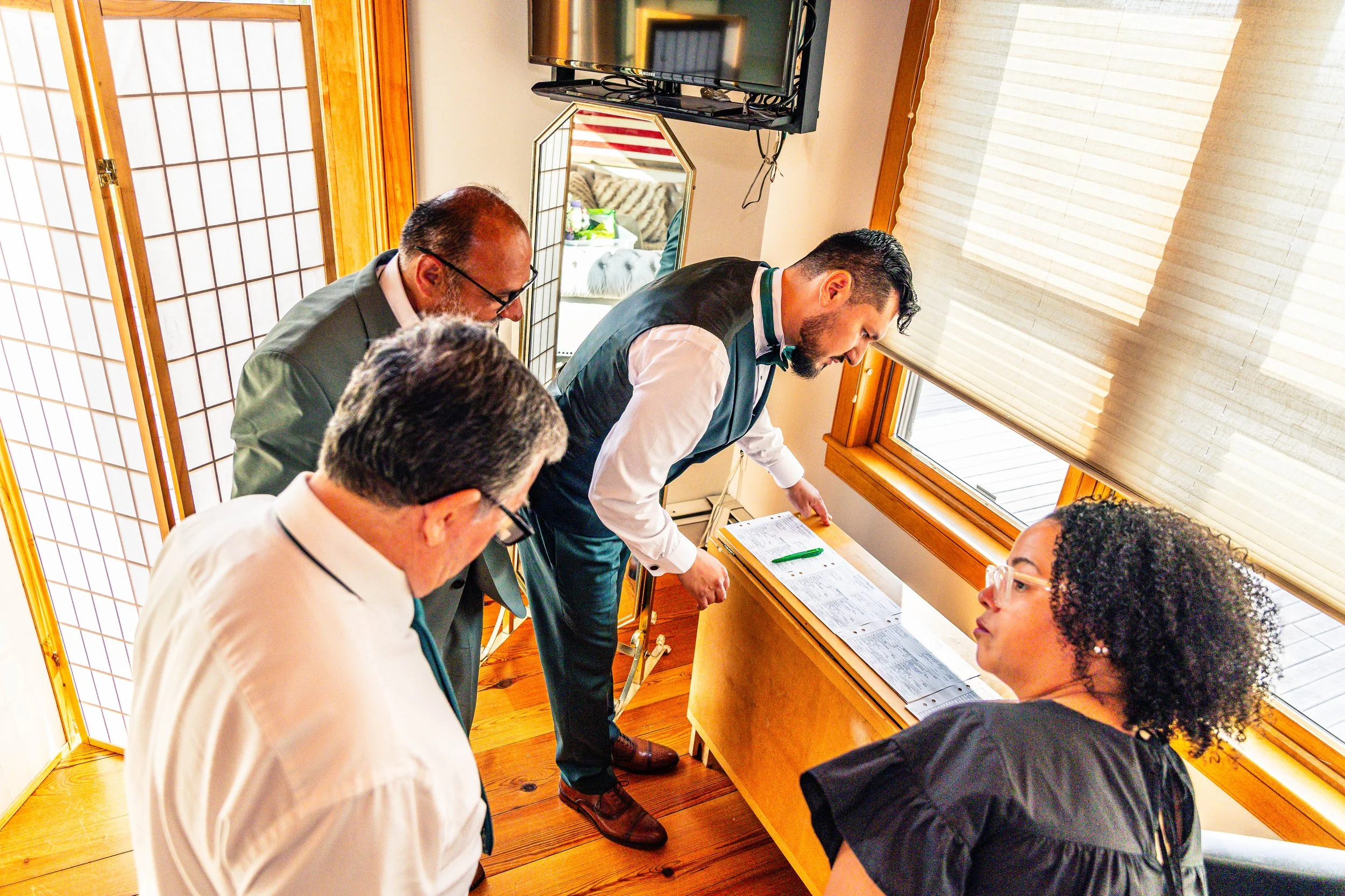 A group of five people gathered around a corner window, reviewing documents on a small table, during a meeting in a sunny room with wooden floors and screens.