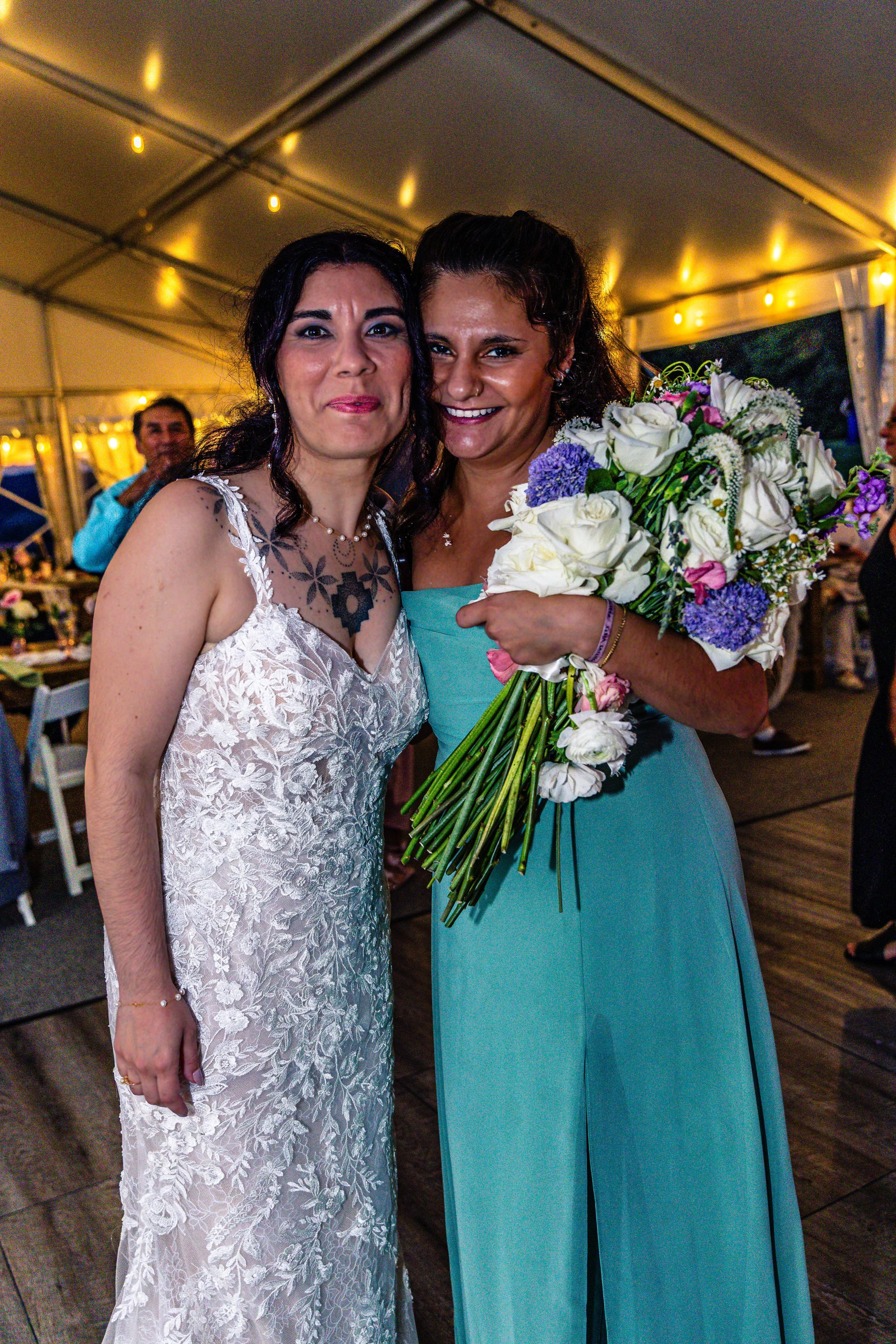 Two women at a celebration, one holding a large bouquet of flowers, smiling and standing closely together in a tent decorated with warm string lights.