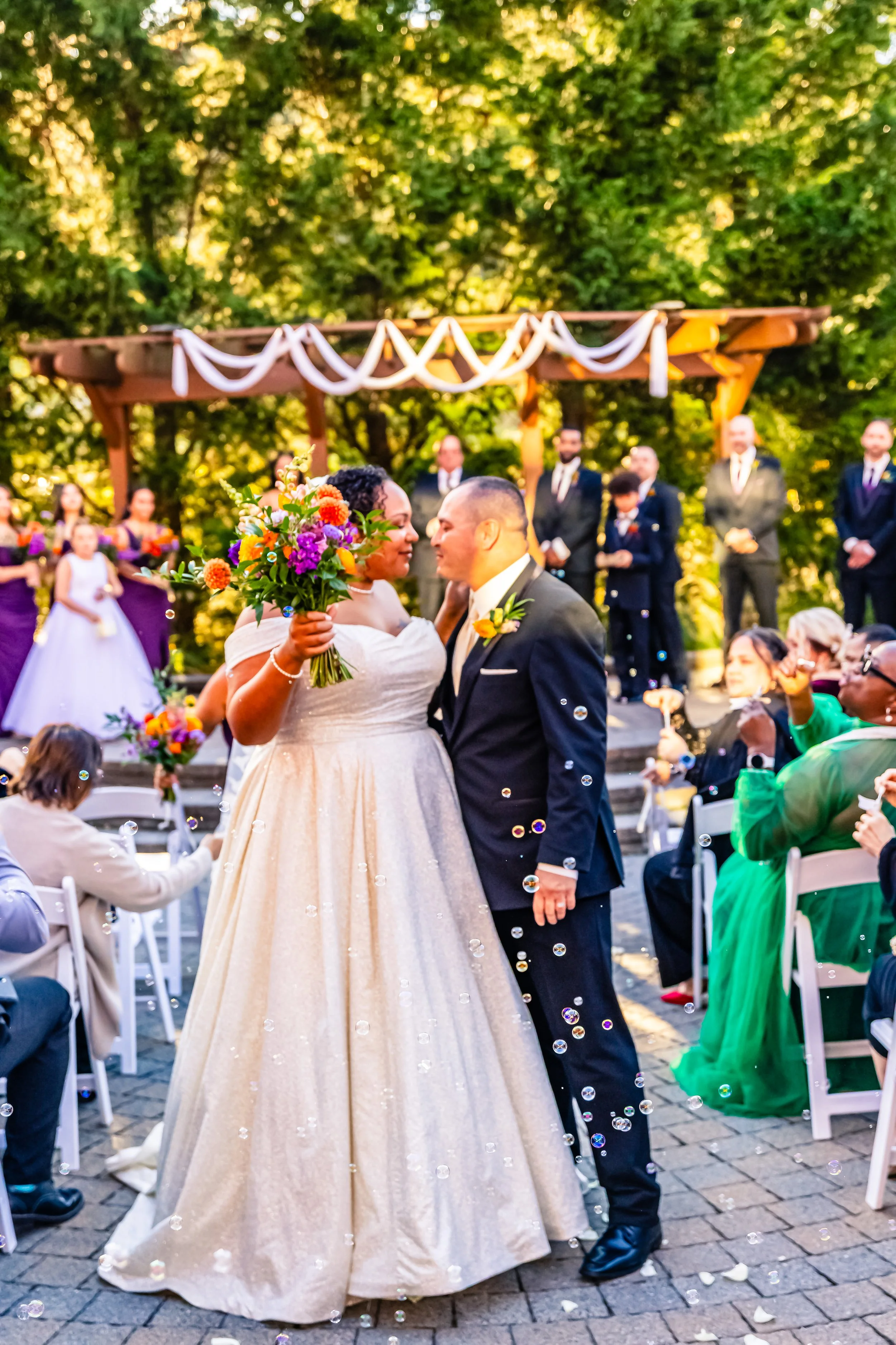 A wedding ceremony outdoors with a bride and groom sharing a moment, surrounded by guests, a wedding party, and floral decorations, with bubbles floating in the air.