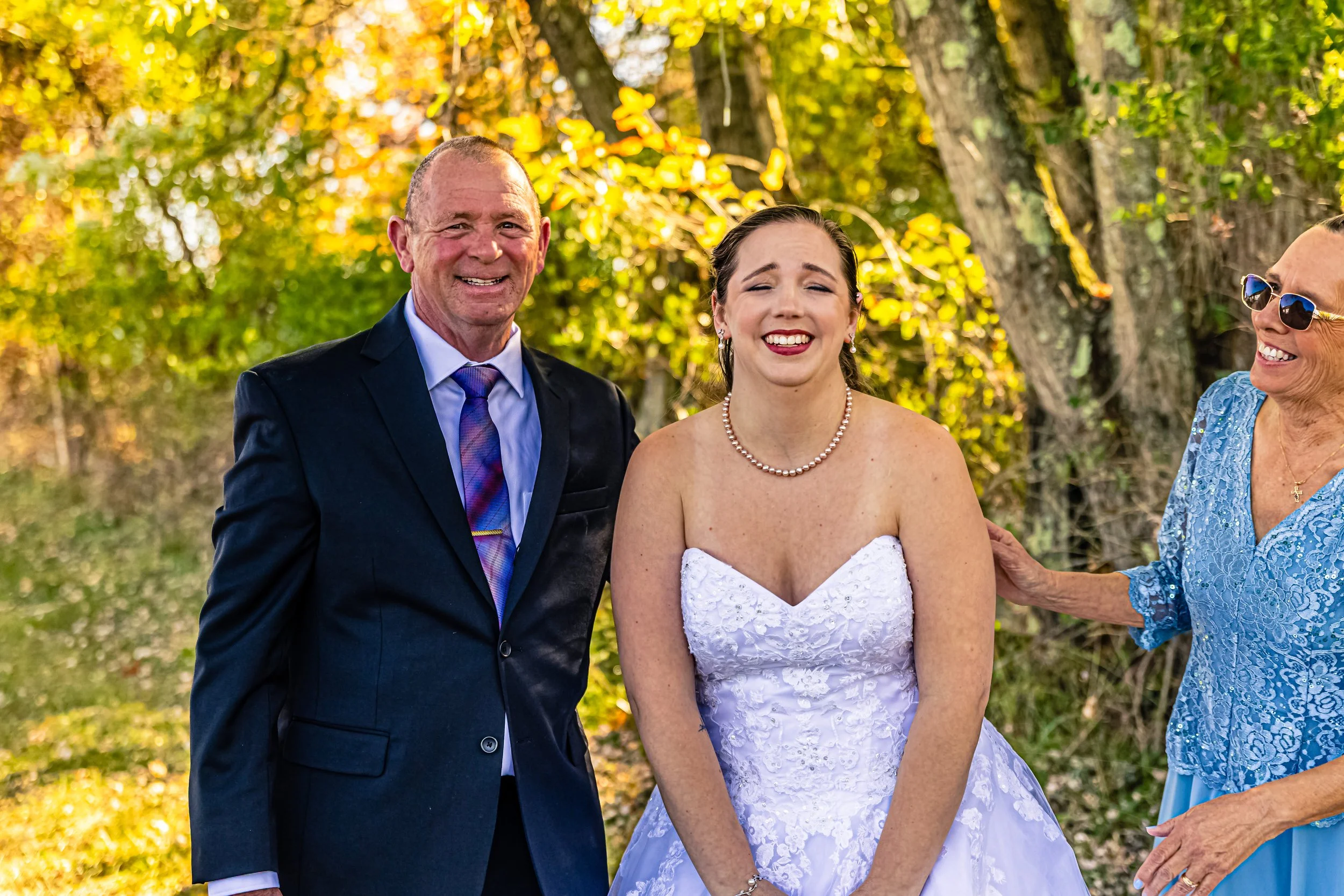 A bride in a white wedding dress and pearl necklace standing next to a smiling man in a navy suit, with an older woman in sunglasses and a blue dress touching her arm, outdoors with autumn foliage in the background.