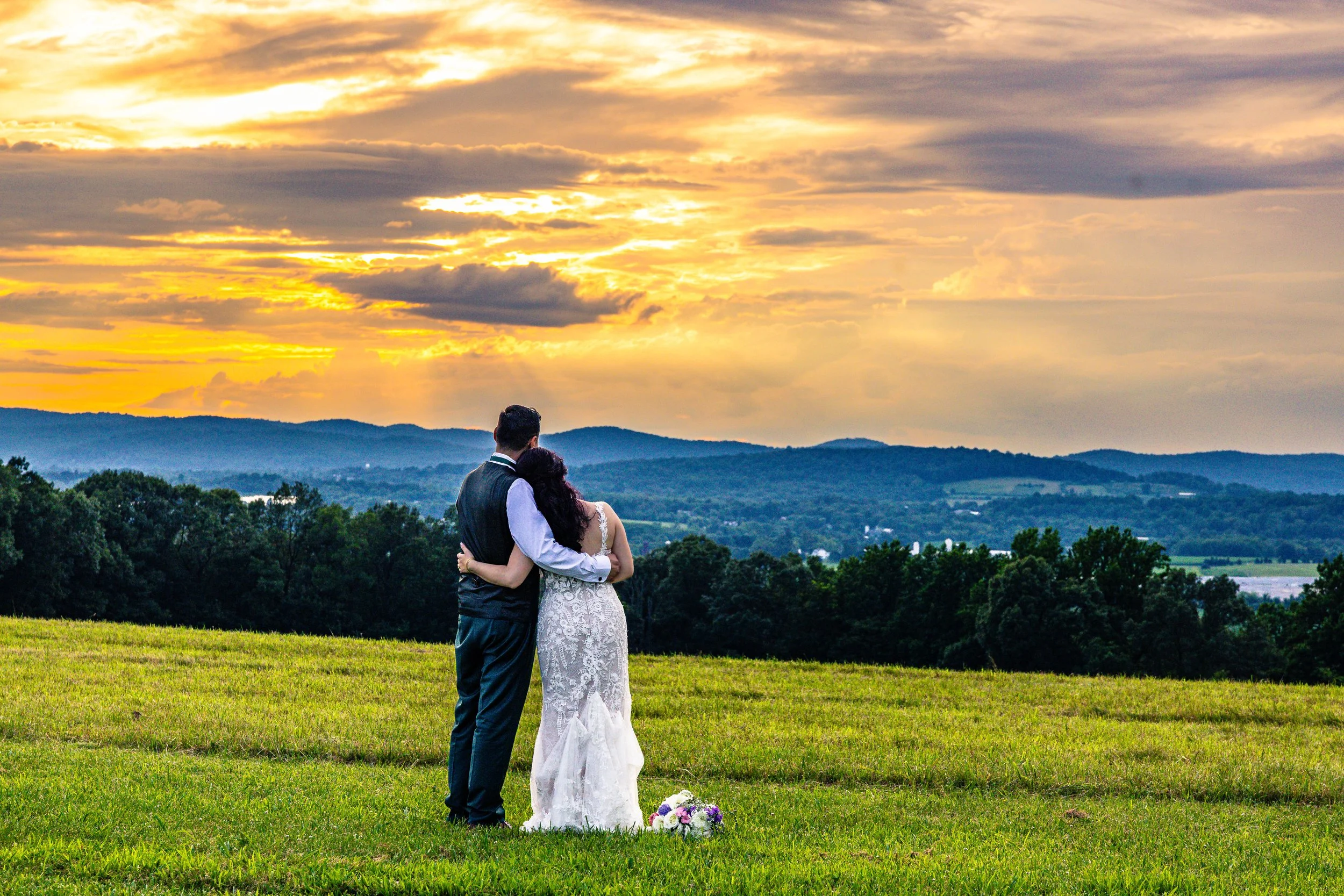 A bride and groom stand closely together on a grassy hill, overlooking a scenic landscape of trees and rolling hills at sunset. The bride is wearing a white lace wedding dress, and the groom is dressed in a dark suit with a white shirt. A bouquet of 