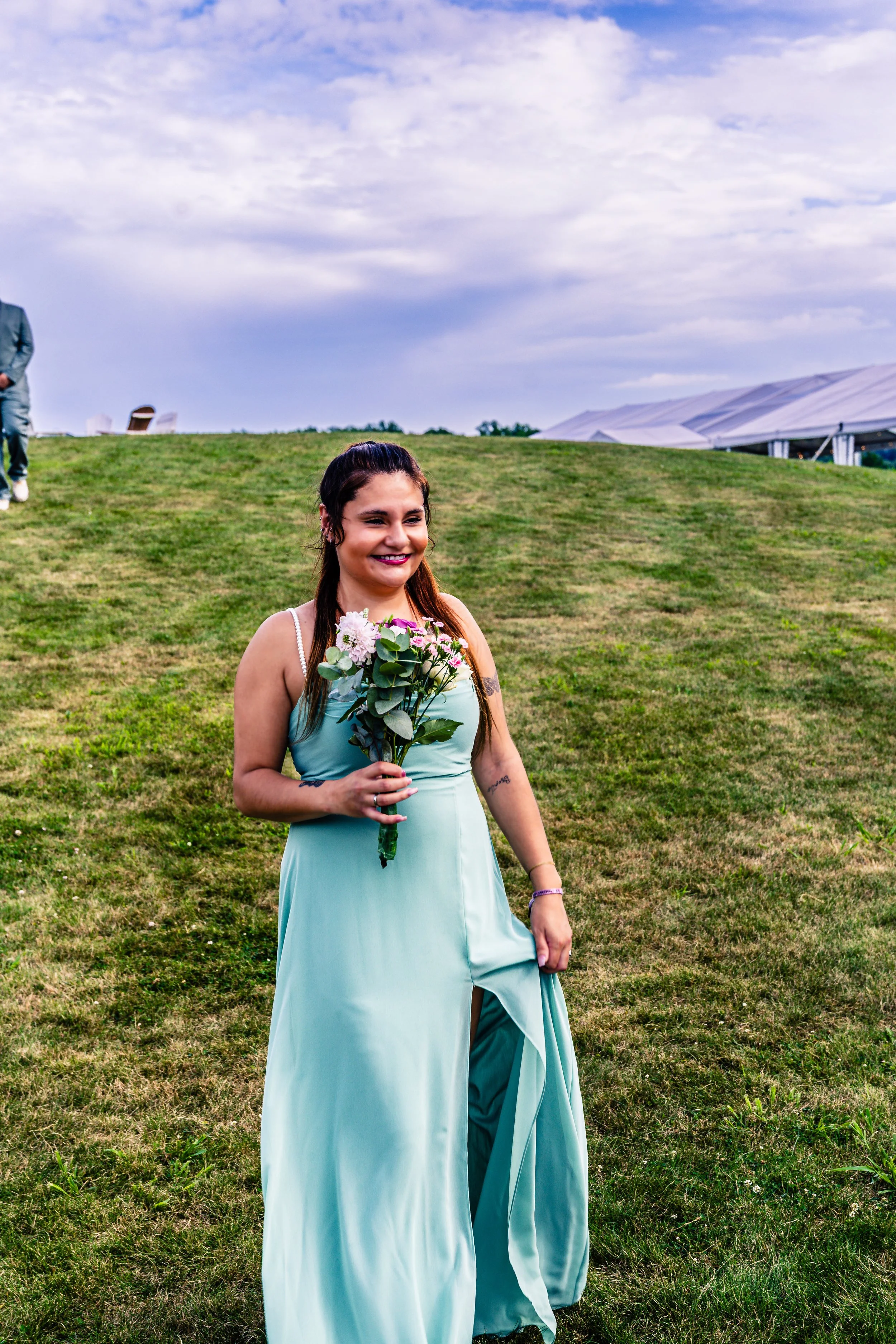 A young woman in a light blue dress holding a bouquet of pink and white flowers on a grassy field, smiling, with a cloudy sky and tents in the background.