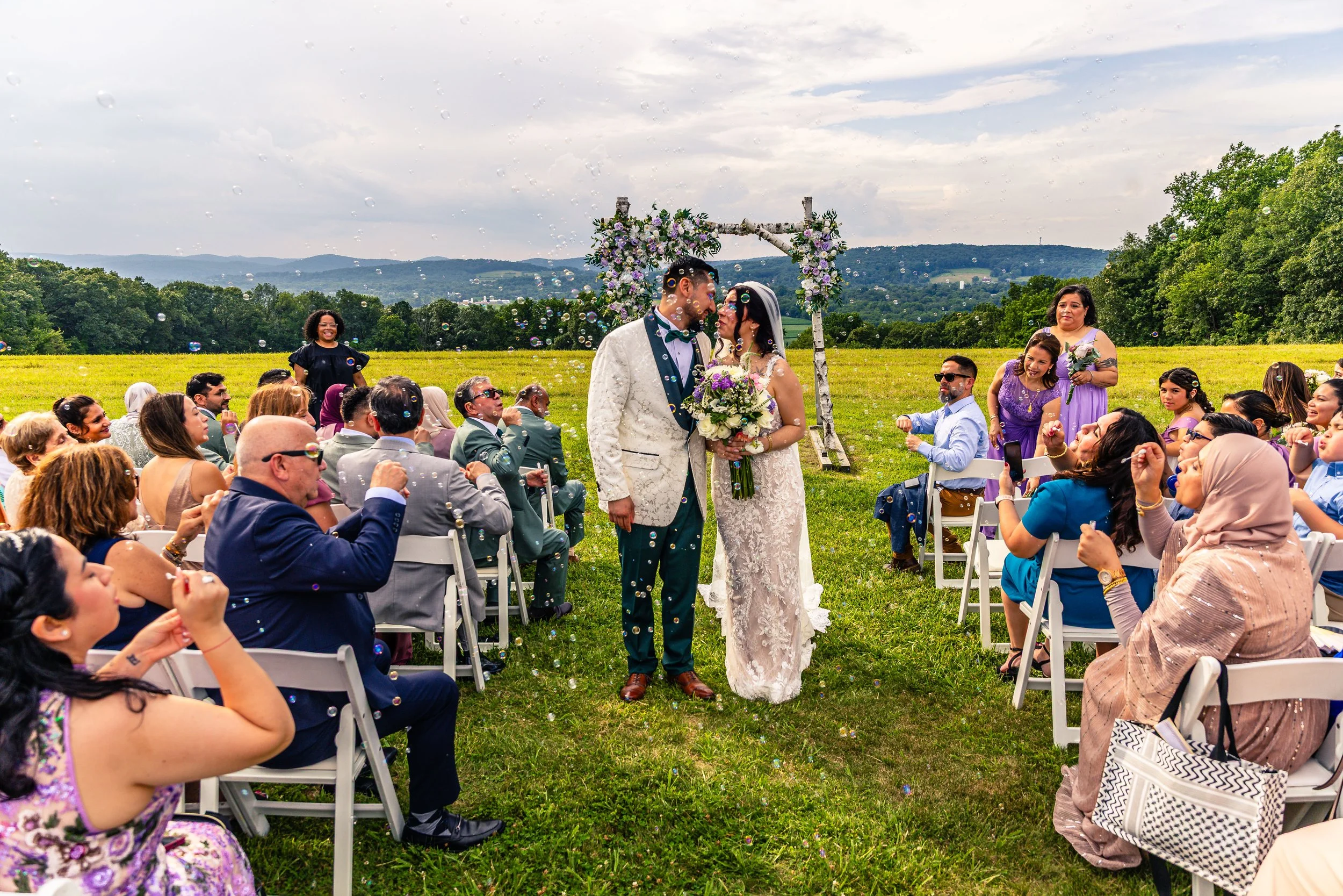 Outdoor wedding ceremony with a bride and groom sharing a kiss, surrounded by seated guests throwing bubbles, on a green field with mountains in the background.