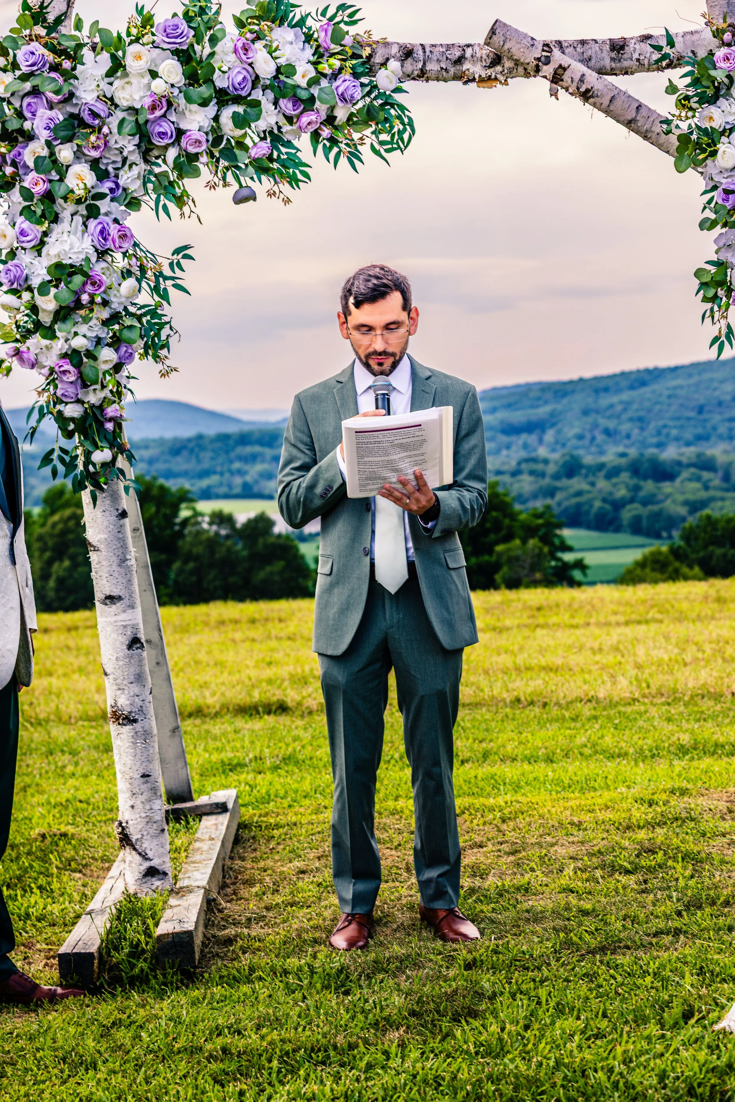 Groom reading vows during outdoor wedding ceremony on lush green field with mountain view, decorated with floral arch of white and purple roses and green foliage.