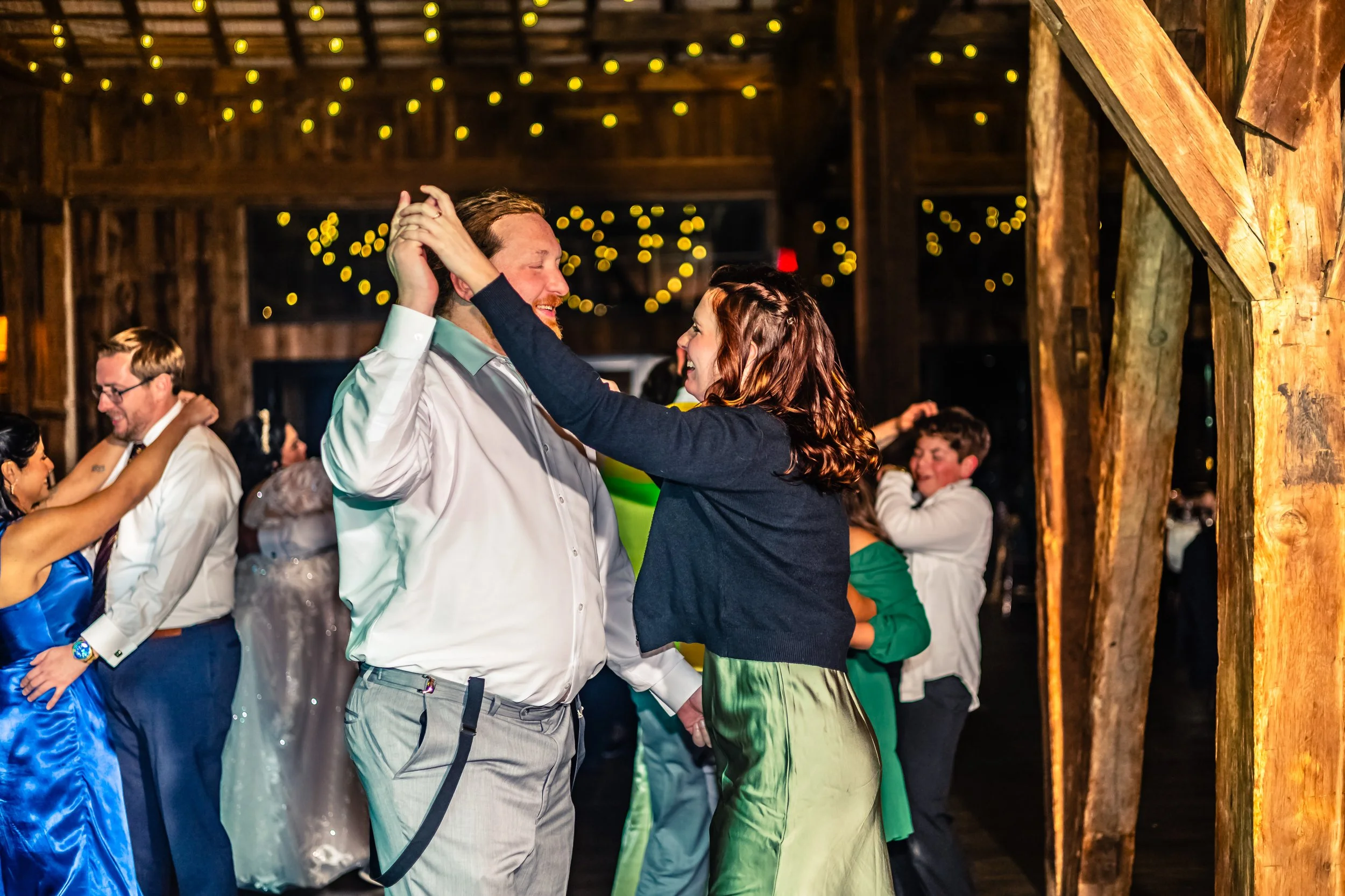 A couple dancing and smiling at a party in a rustic barn, with string lights hanging from the ceiling and other guests dancing in the background.