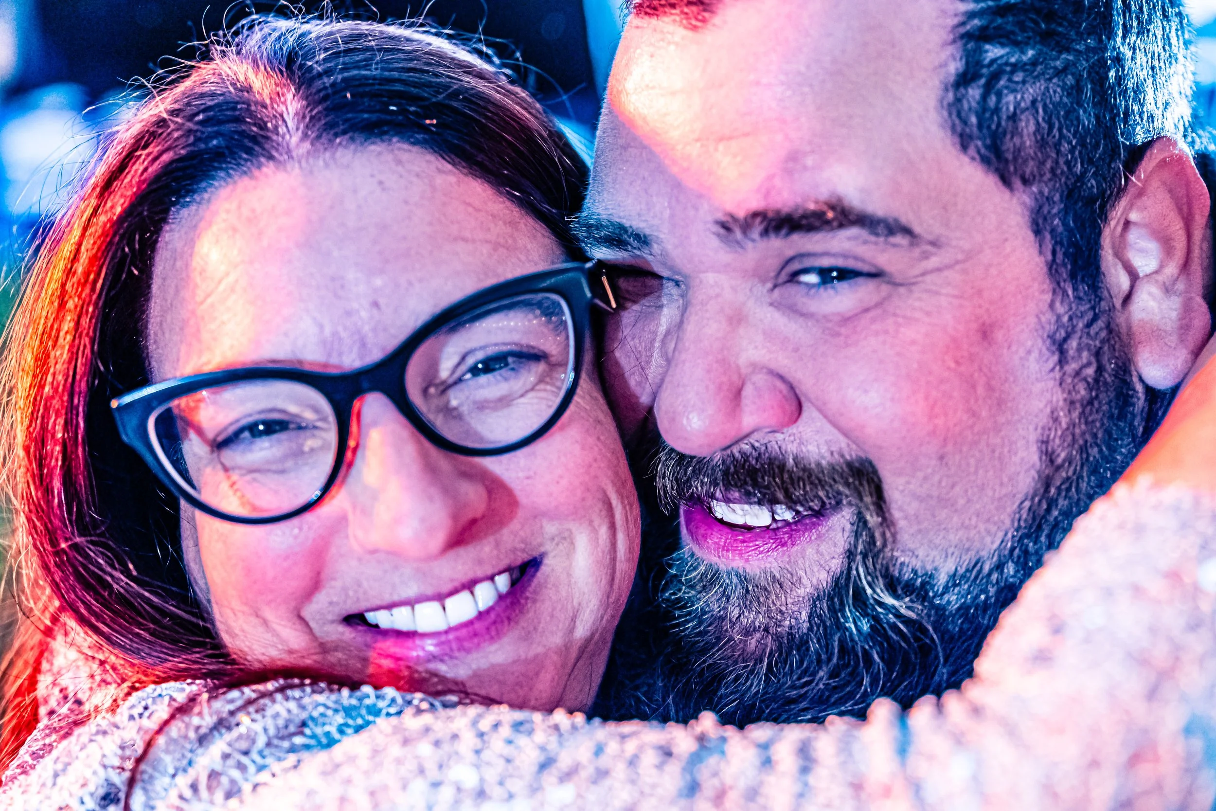 Close-up of a smiling woman with glasses and a smiling man hugging in bright outdoor setting.