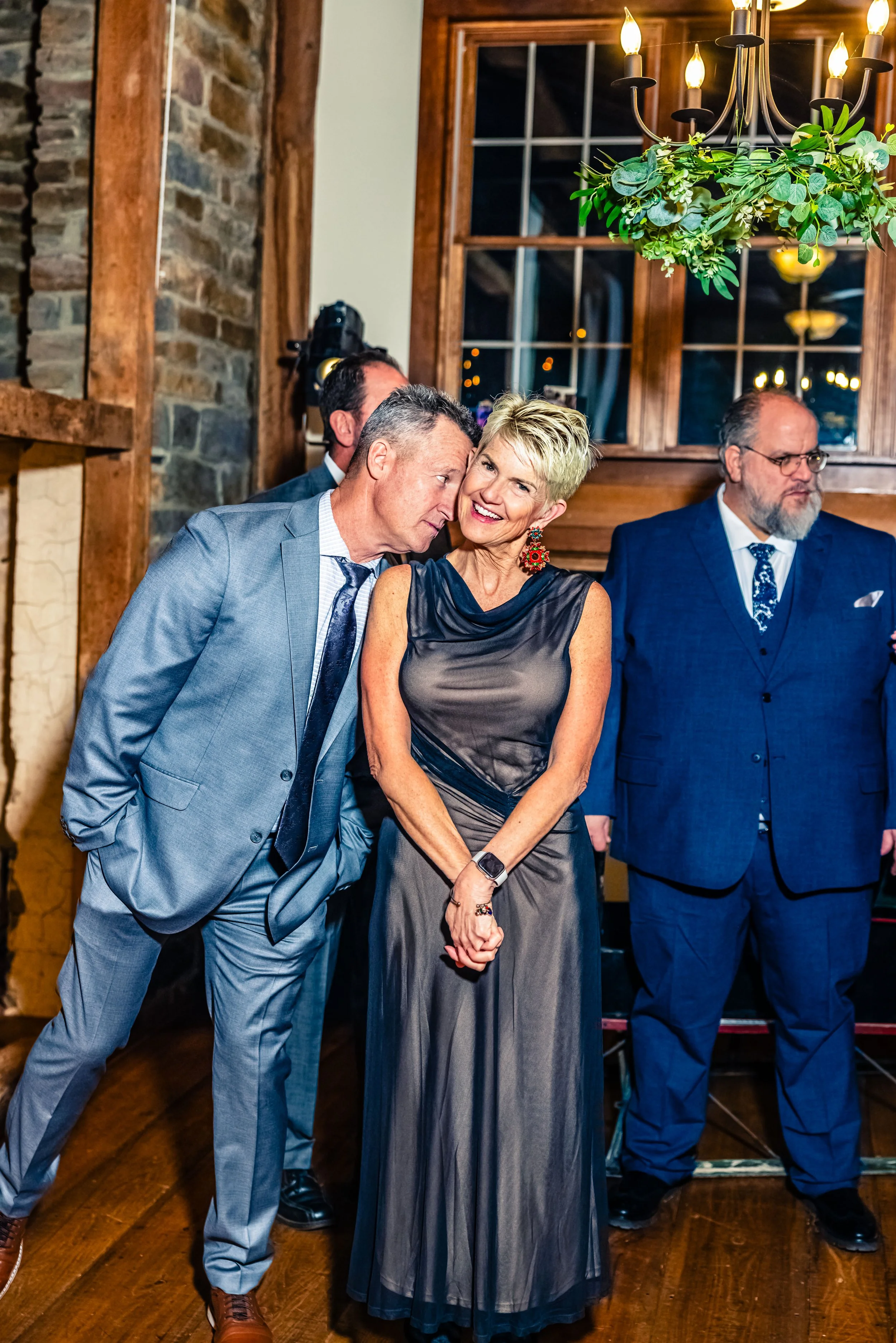 A man whispering to a woman at a social event in a warm, rustic indoor setting, with other men in suits standing nearby.