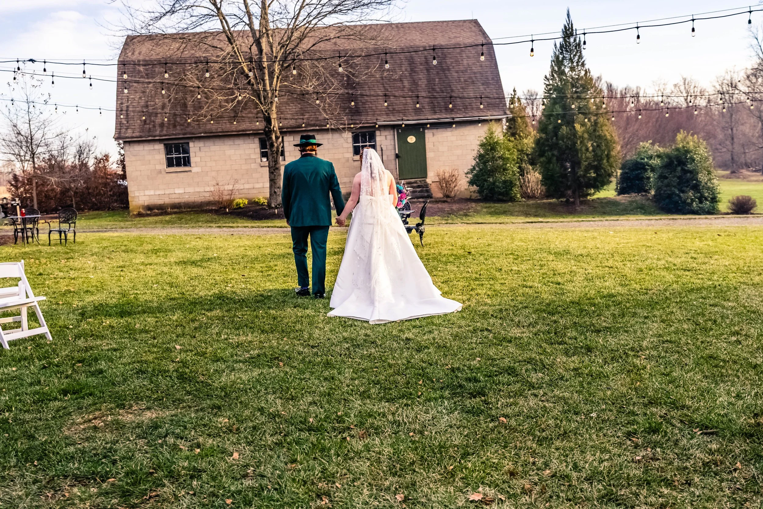 A newlywed couple walking hand-in-hand towards a rustic barn on a grassy field, with string lights hanging above them and a parked bicycle nearby.