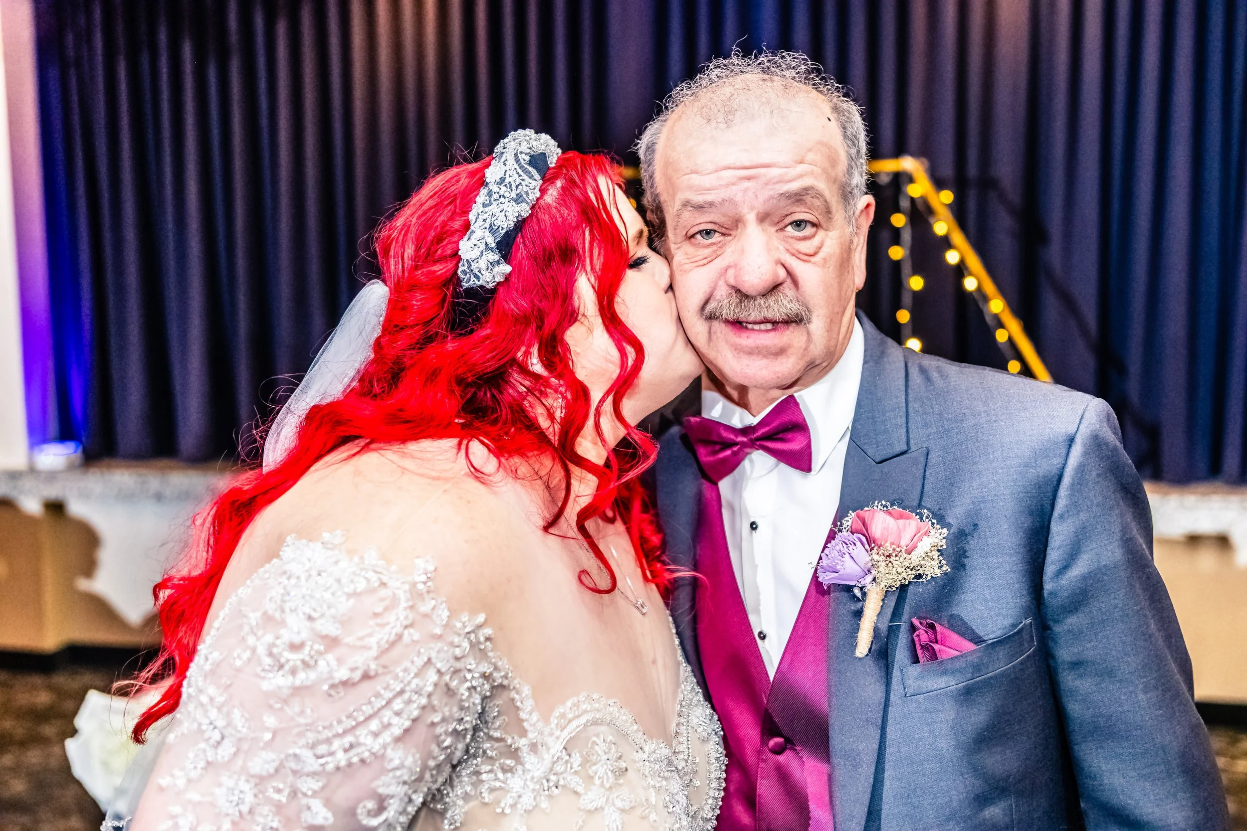 A bride with bright red hair, wearing a white lace wedding dress, kisses an older man with a mustache, dressed in a blue suit with a purple bow tie, on a wedding stage.