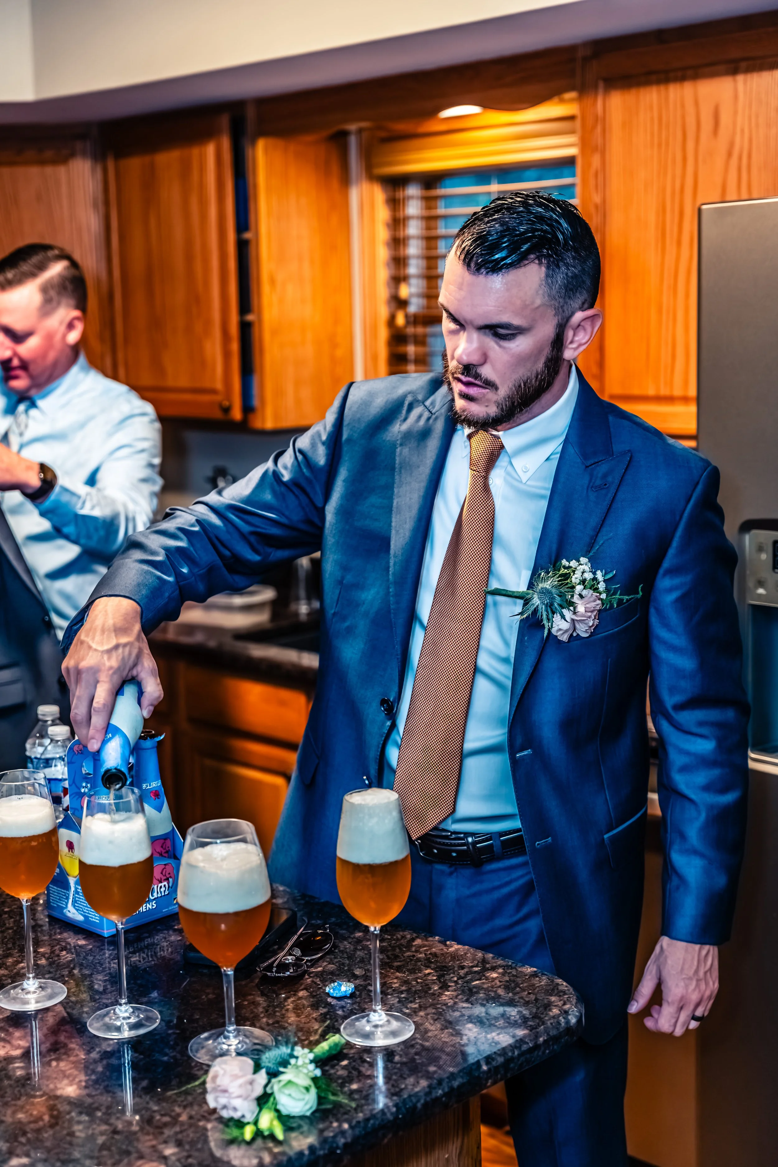 A man in a blue suit and a boutonniere pours drinks at a celebration, with others in the background in a kitchen setting.