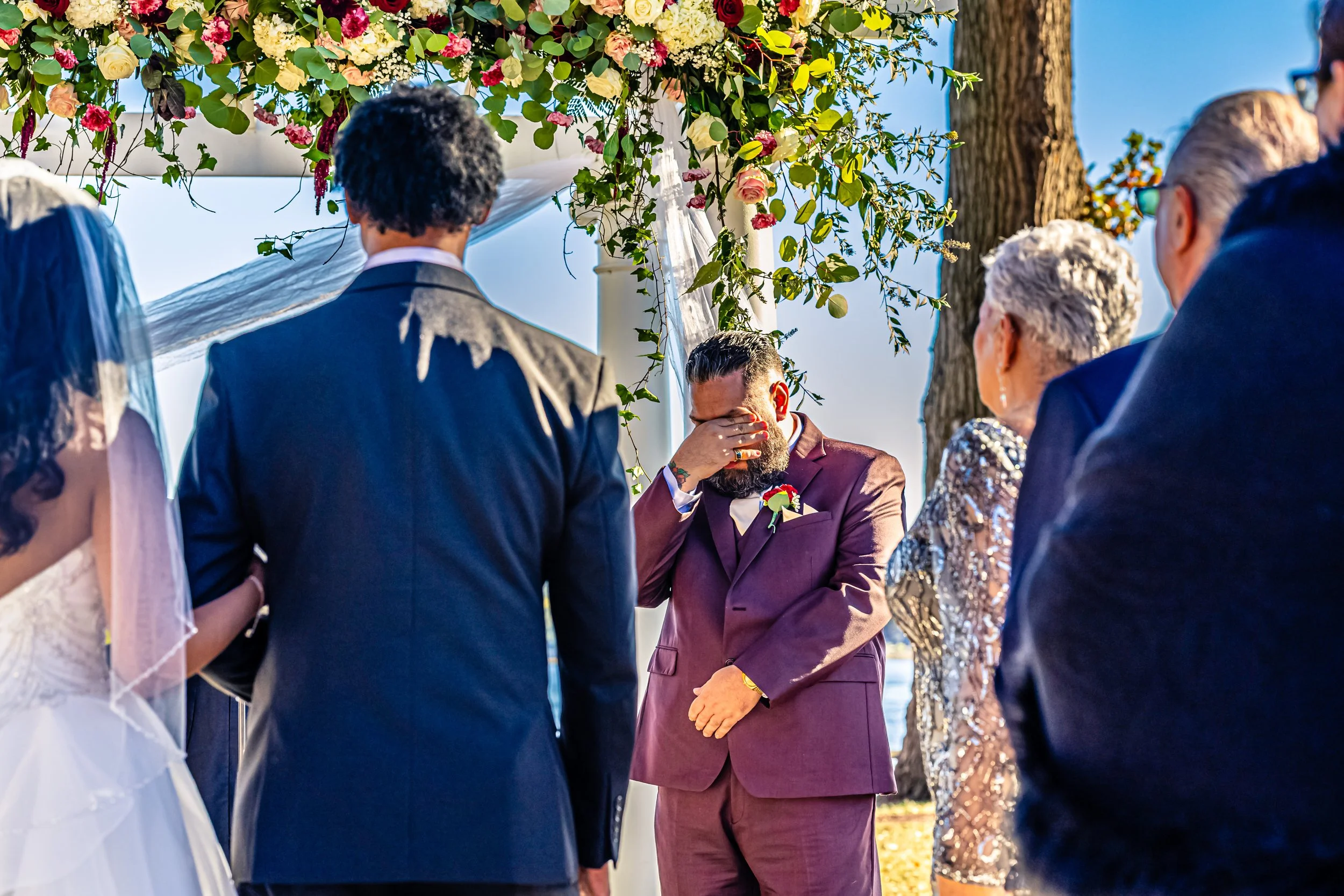 A wedding ceremony outdoors with a groom in a maroon suit covering his face, facing a bride and guests under a decorated archway with flowers and greenery.