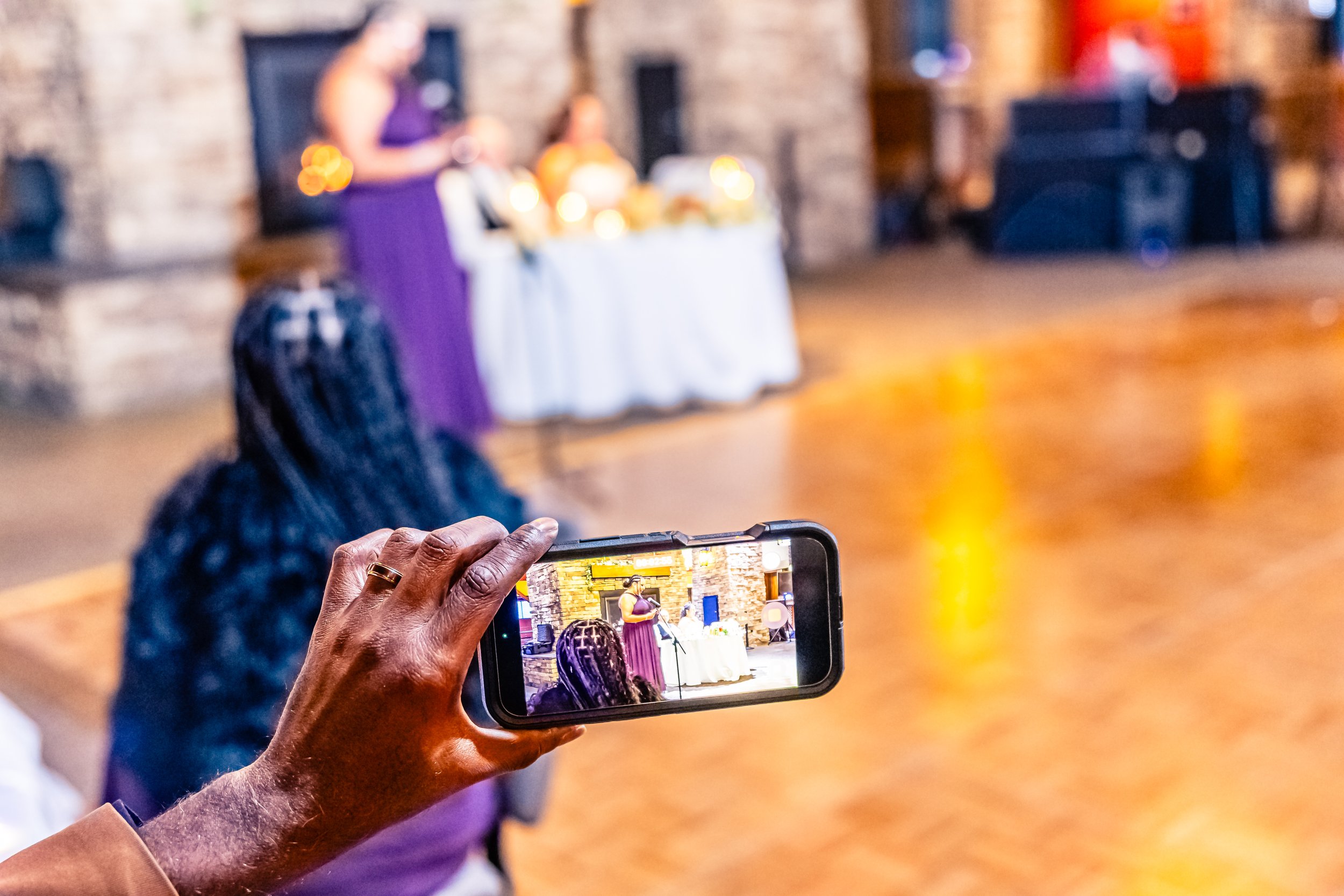 Person taking a photo of a woman speaking at a microphone during an event in a rustic indoor venue with brick walls and a white tablecloth.