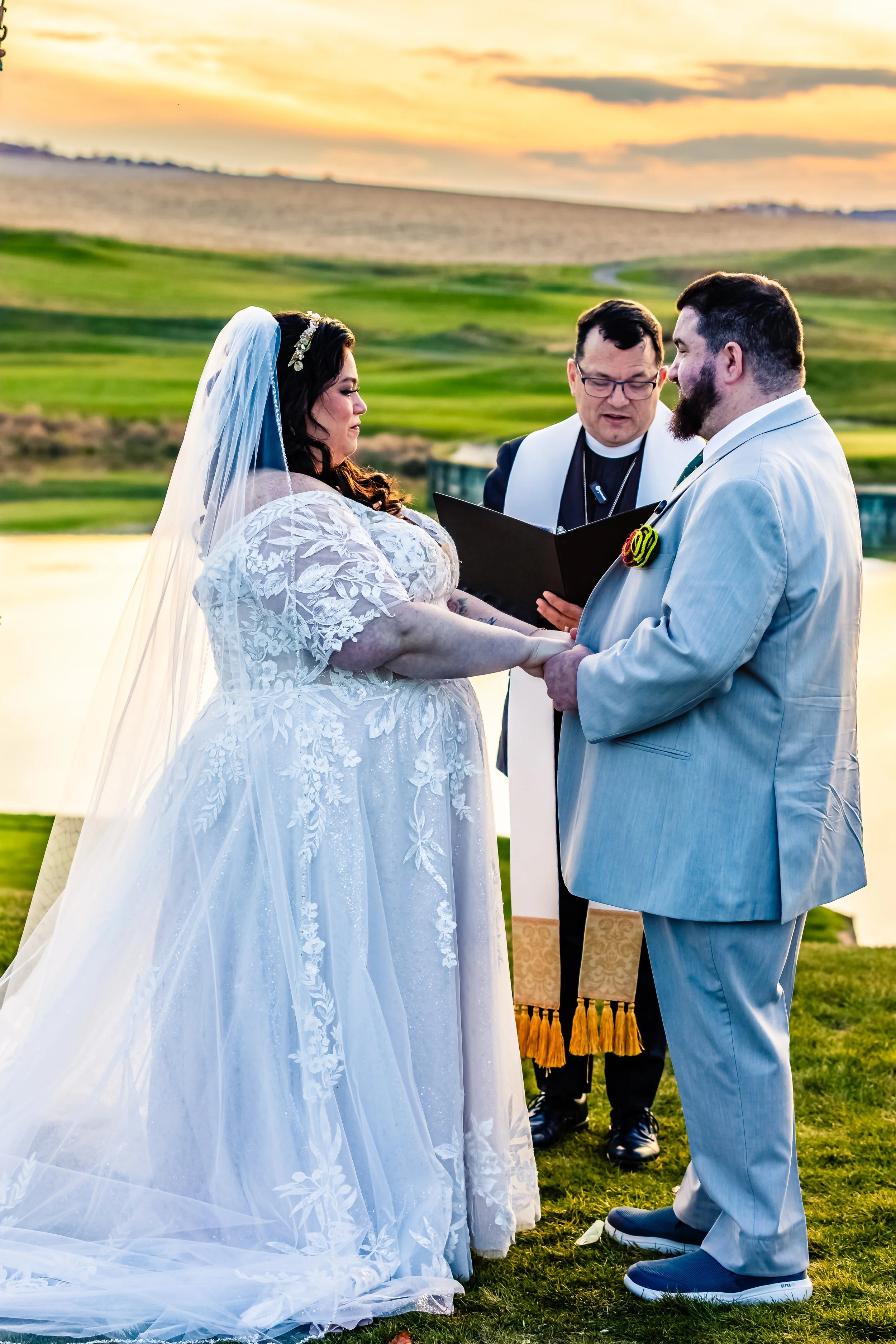 A couple getting married outdoors by a lake with a sunset sky, holding hands and looking at each other, with an officiant reading from a book in front of them.