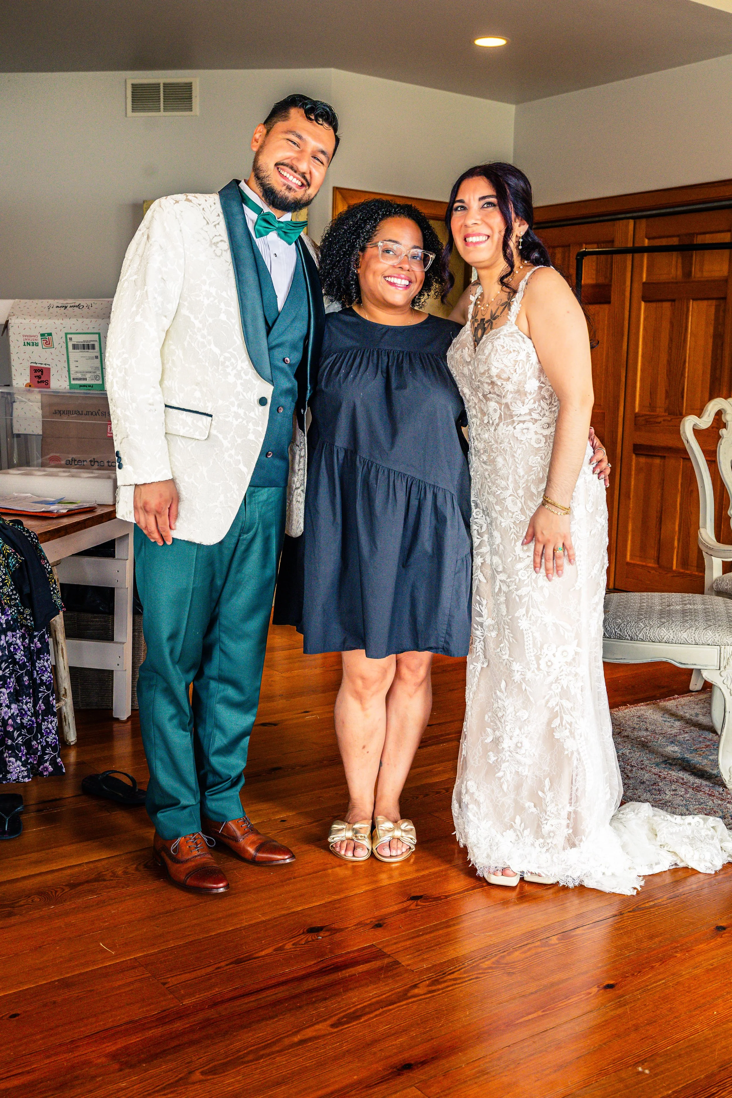 Three people smiling at a celebration, with a man in a tuxedo with a white jacket, a woman in a wedding dress, and another woman in a dark blue dress, standing on a wooden floor indoors.