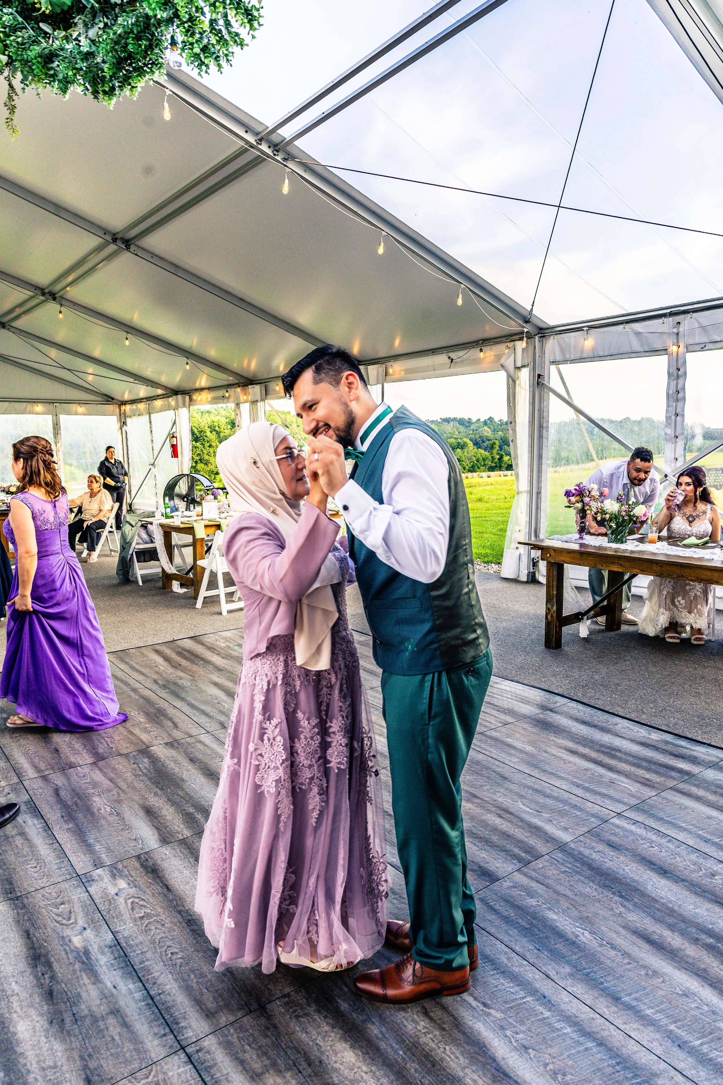 A young man and an older woman are dancing and smiling at a wedding reception under a large outdoor tent, with other guests in the background.